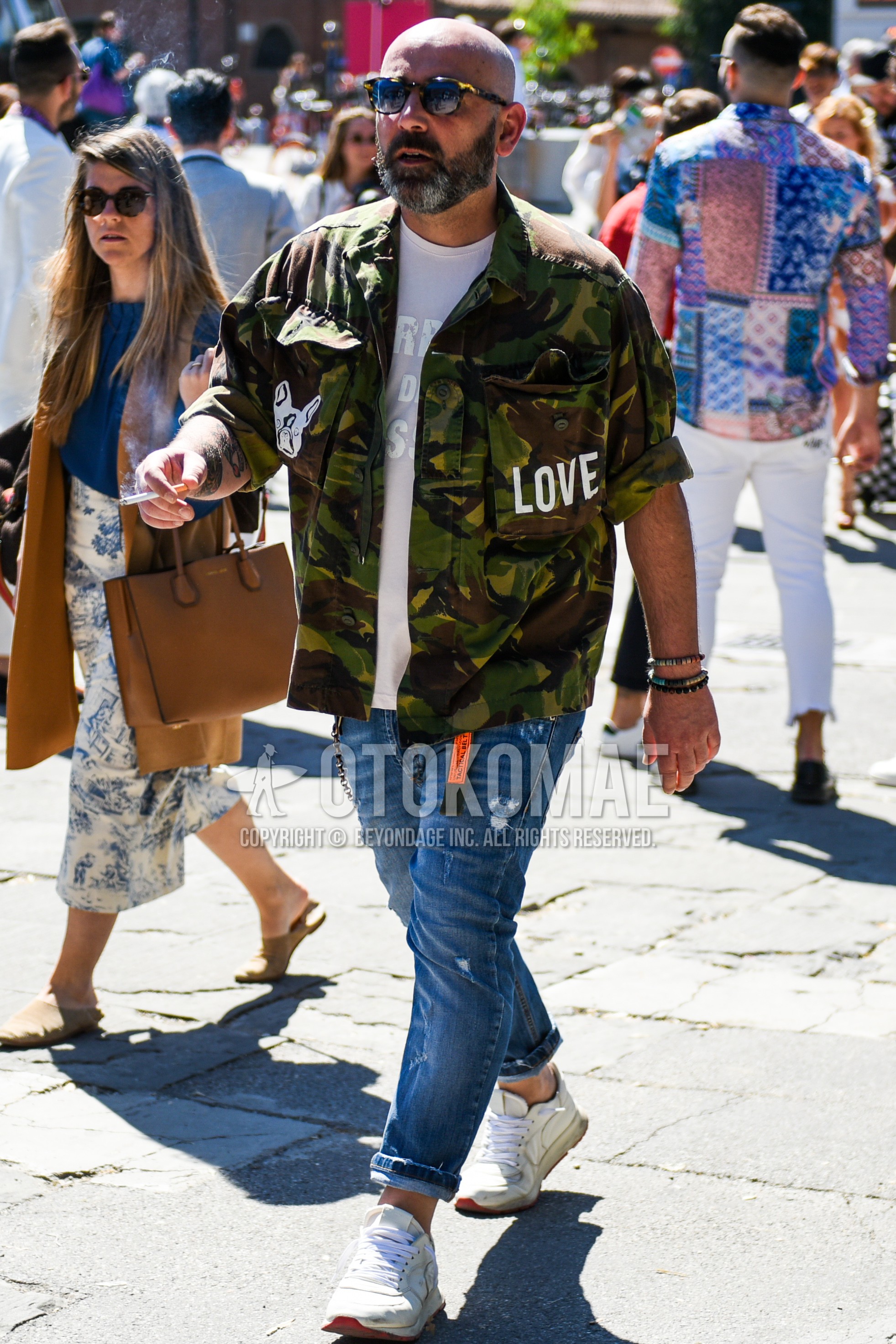 Men's spring autumn outfit with beige tortoiseshell sunglasses, olive green camouflage shirt jacket, white graphic t-shirt, blue plain damaged jeans, white low-cut sneakers.