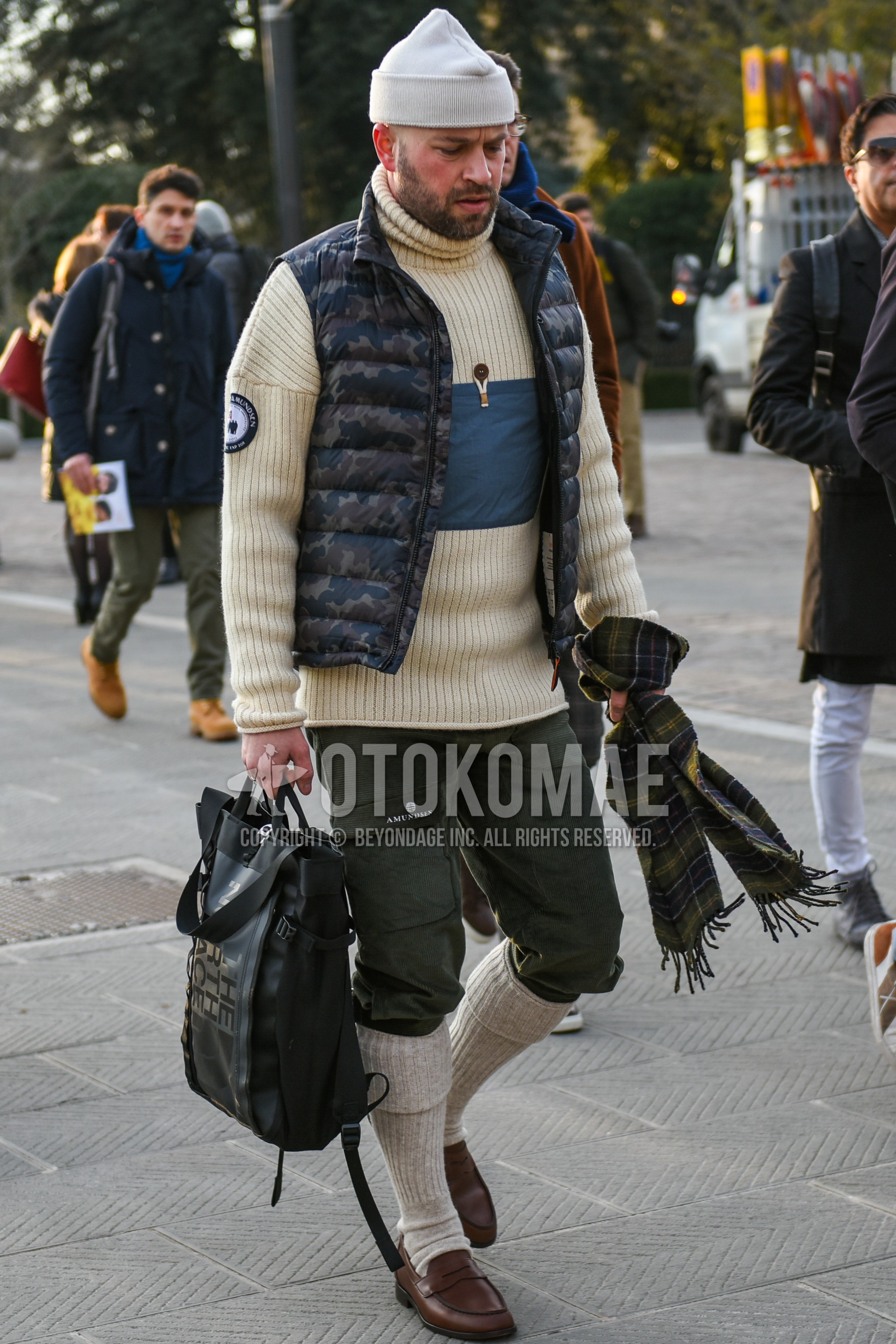 Men's autumn winter outfit with white plain knit cap, green camouflage down jacket, olive green plain cargo pants, beige plain socks, brown coin loafers leather shoes, green camouflage briefcase/handbag.