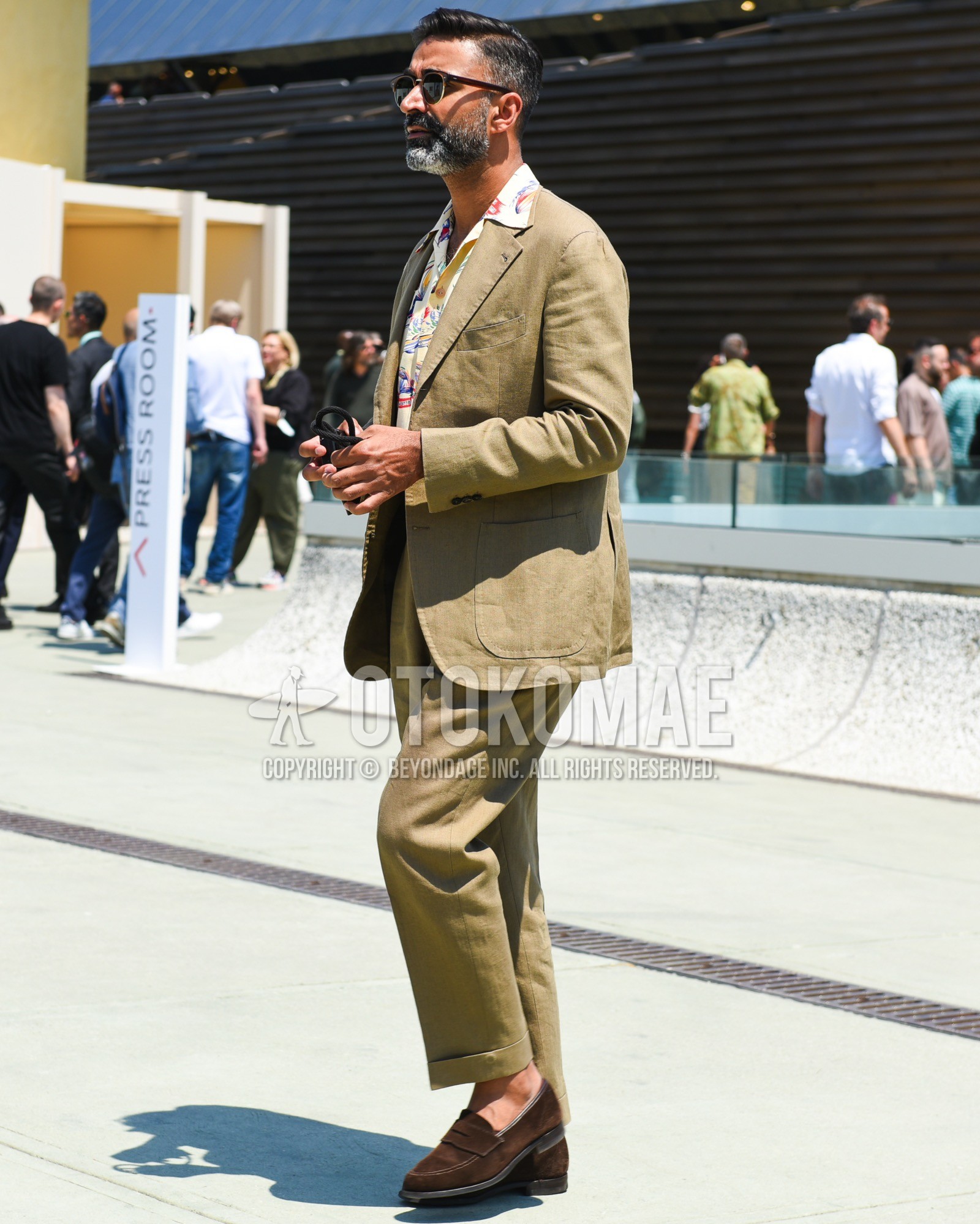 Men's spring summer outfit with brown tortoiseshell sunglasses, white graphic shirt, brown coin loafers leather shoes, beige plain suit.