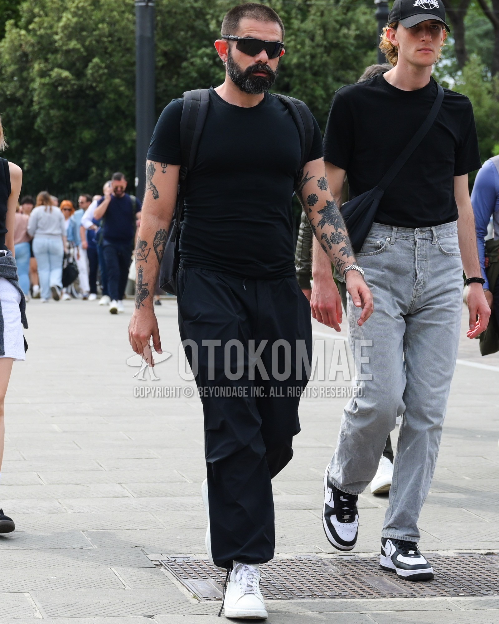 Men's spring summer outfit with black plain sunglasses, black plain t-shirt, black plain wide pants, white low-cut sneakers, black plain backpack.