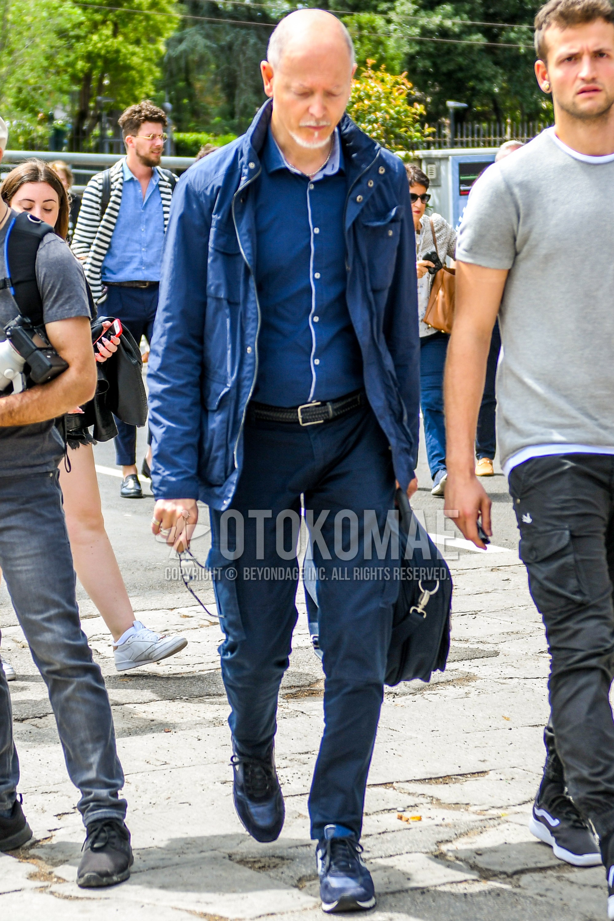 Men's spring autumn outfit with navy plain M-65, navy plain shirt, black plain leather belt, black plain braided belt, navy plain cargo pants, navy plain chinos, navy low-cut sneakers, navy plain briefcase/handbag.