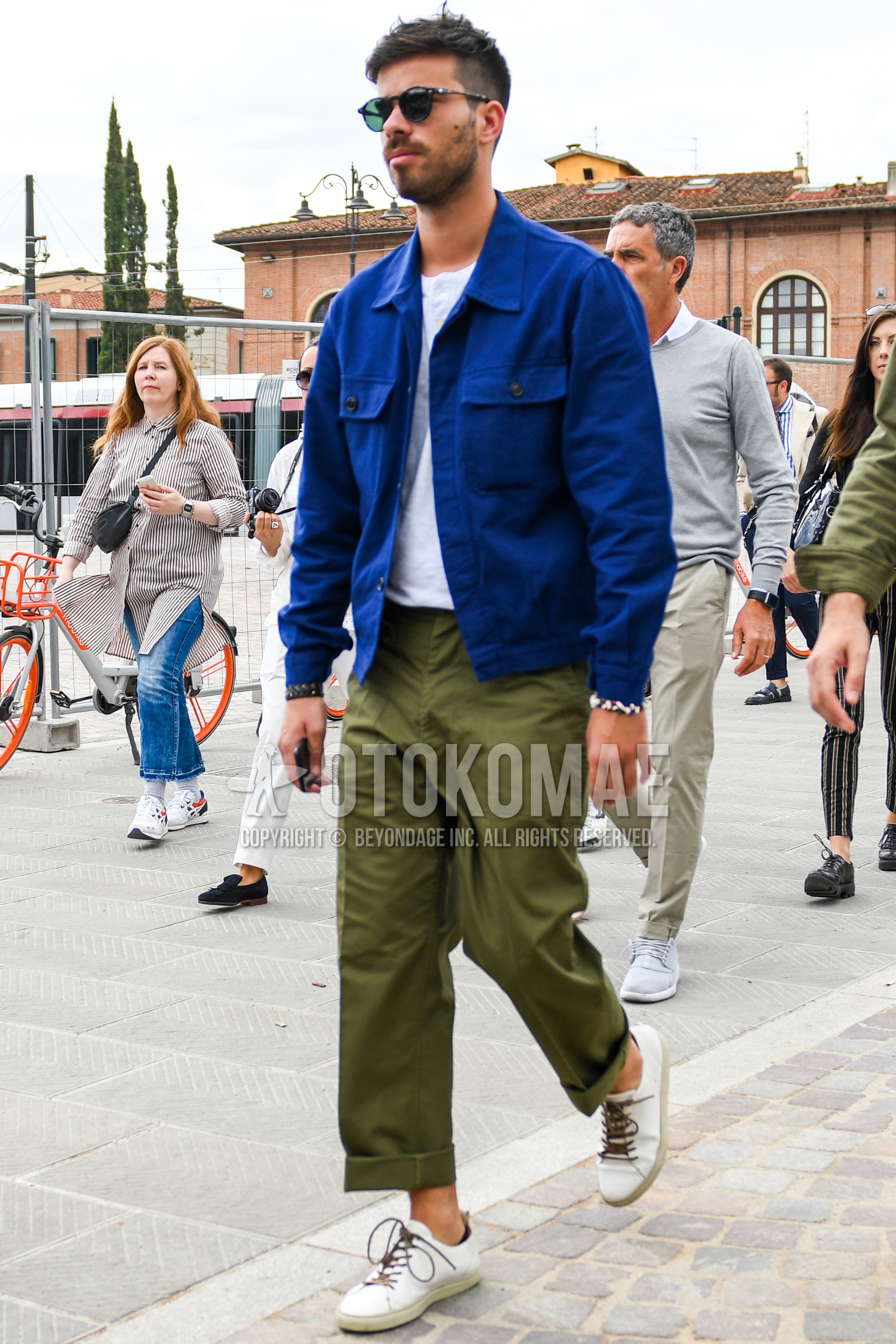 Men's spring summer autumn outfit with plain sunglasses, blue plain trucker jacket, white plain t-shirt, olive green plain chinos, white low-cut sneakers.