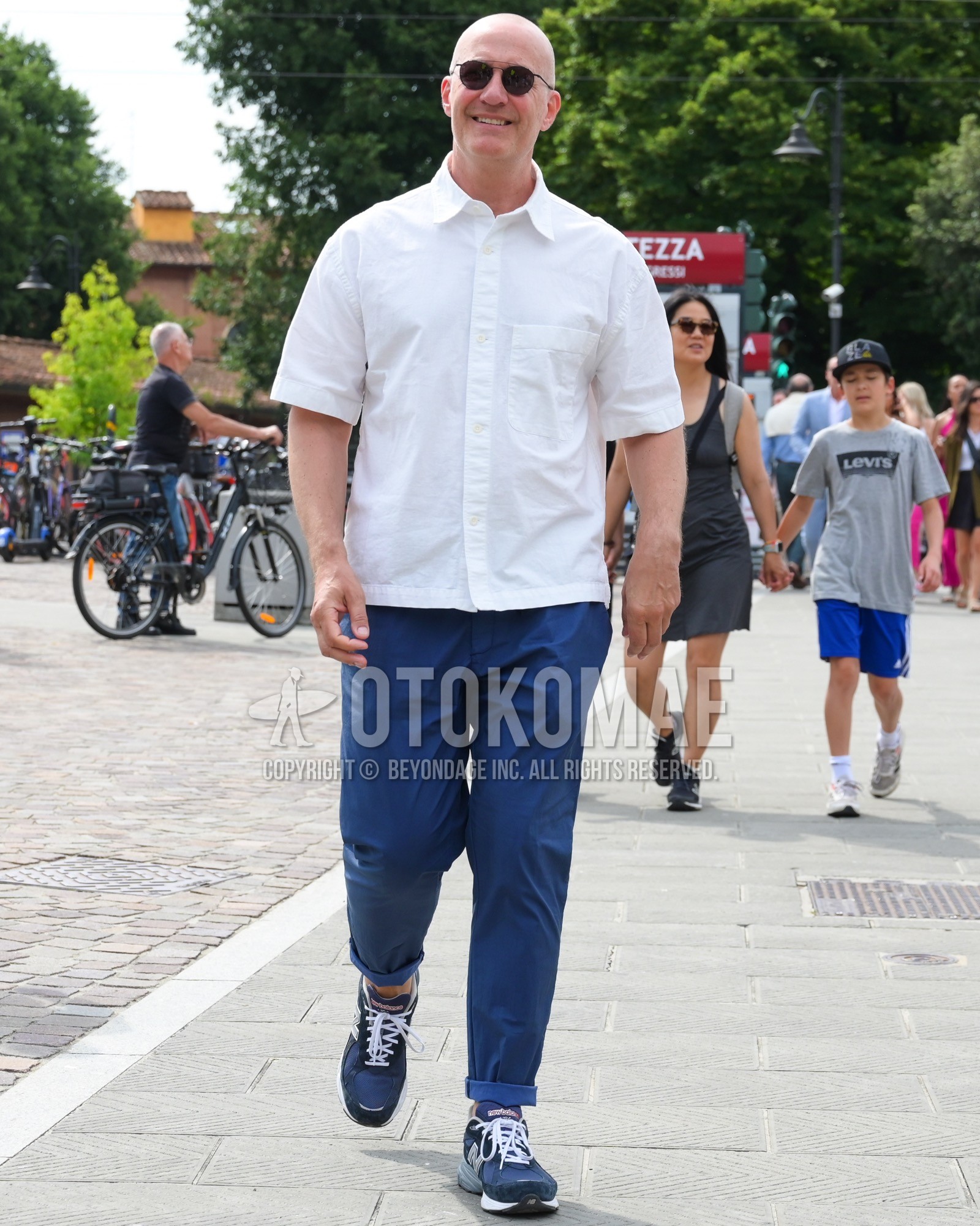 Men's spring summer outfit with black plain sunglasses, white plain shirt, blue plain chinos, blue low-cut sneakers.