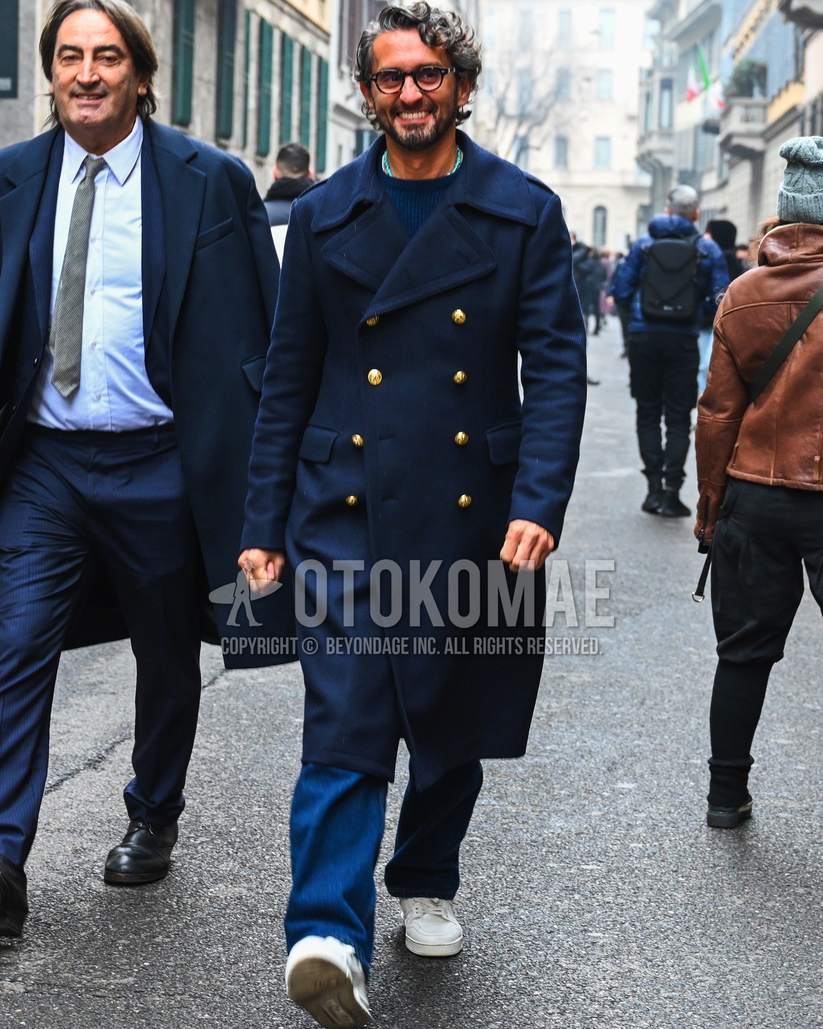 Men's autumn winter outfit with brown tortoiseshell glasses, navy plain ulster coat, navy plain sweater, blue plain denim/jeans, white low-cut sneakers.