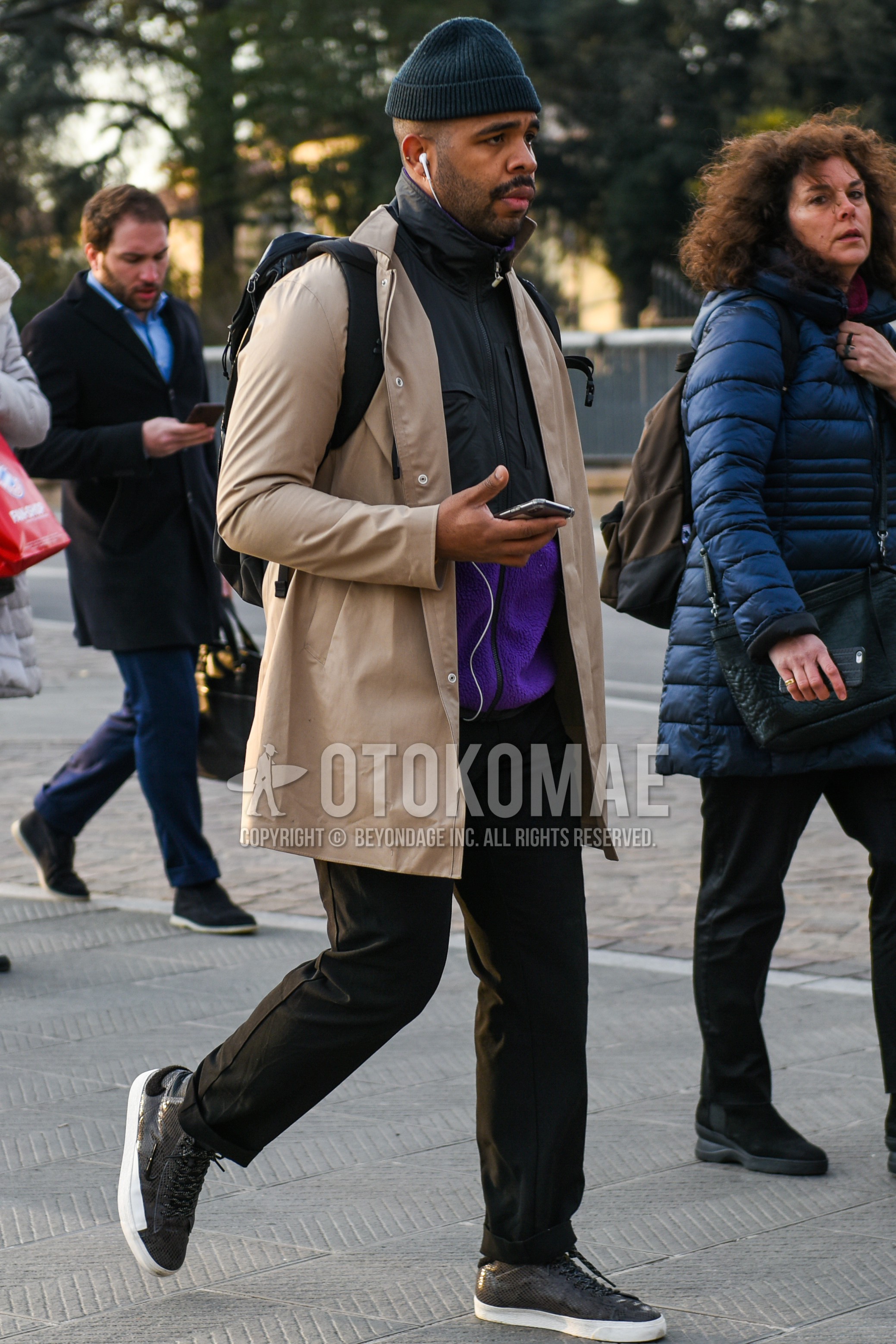 Men's autumn winter outfit with gray plain knit cap, beige plain stenkarrer coat, gray purple plain windbreaker, gray purple plain fleece jacket, black plain slacks, gray low-cut sneakers, black plain backpack.