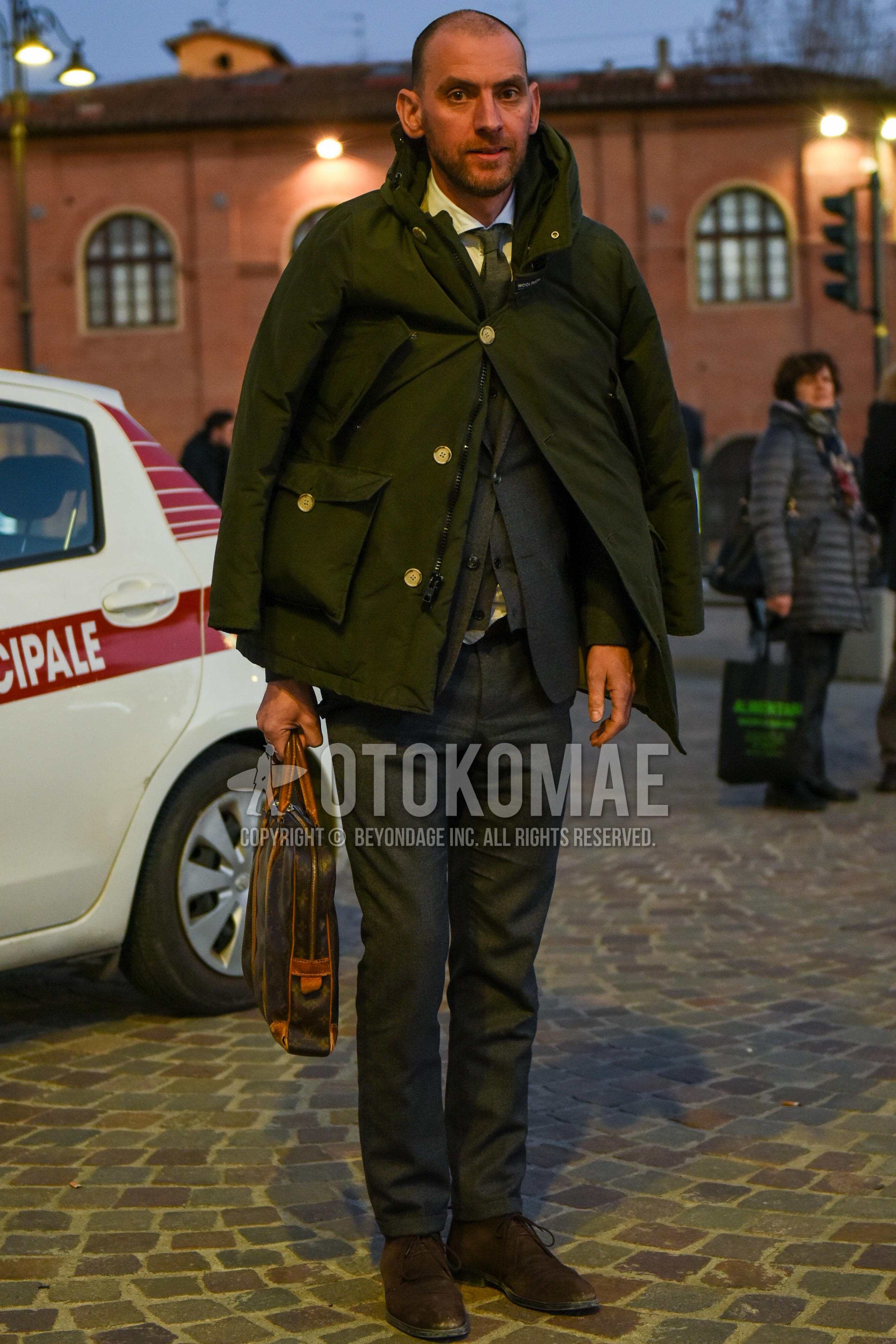 Men's autumn winter outfit with olive green plain down jacket, white plain shirt, gray plain cardigan, brown chukka boots, brown bag briefcase/handbag, gray plain suit, gray plain necktie.