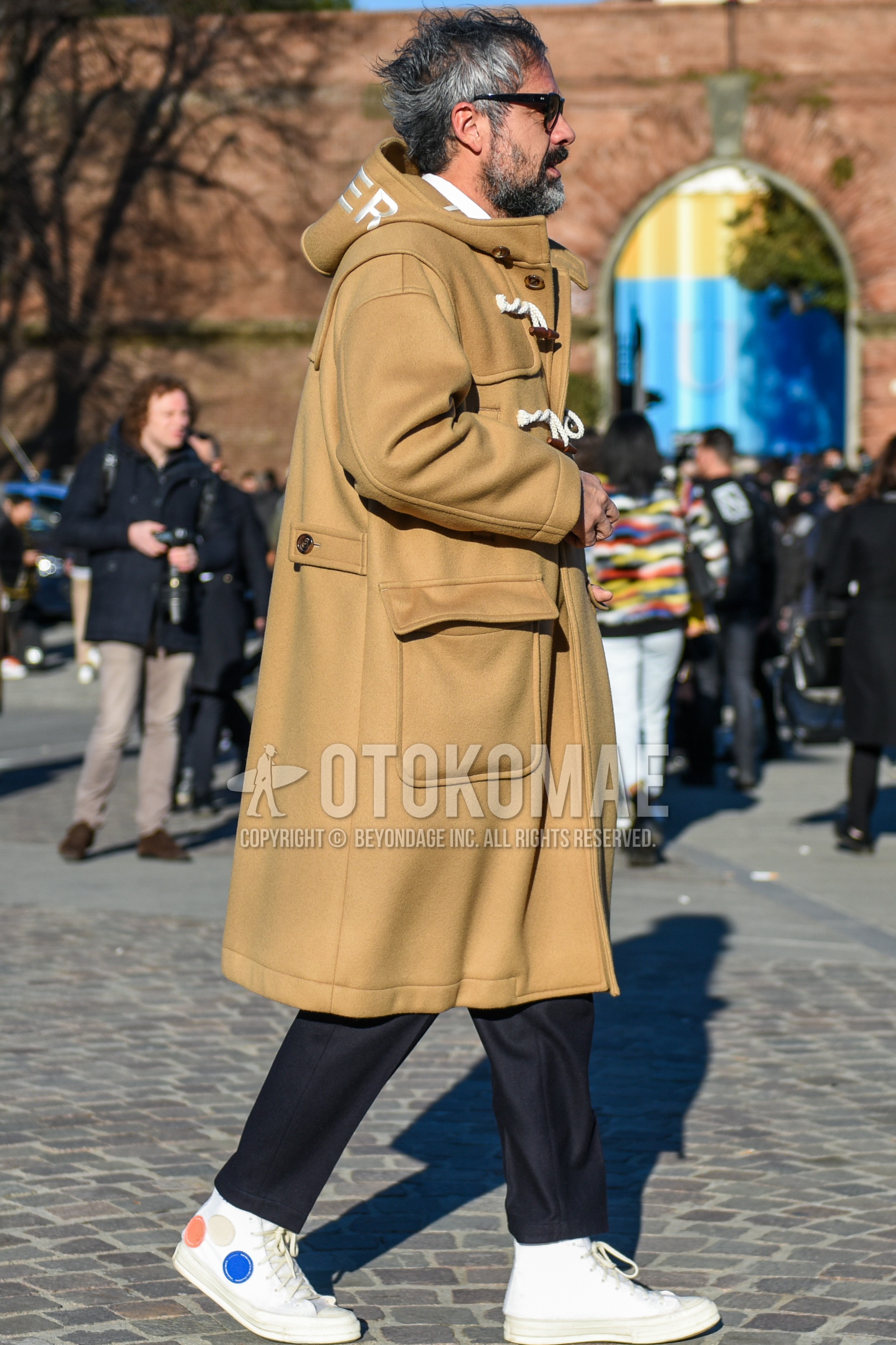 Men's autumn winter outfit with black plain sunglasses, beige plain duffle coat, navy plain slacks, navy plain cropped pants, white high-cut sneakers.