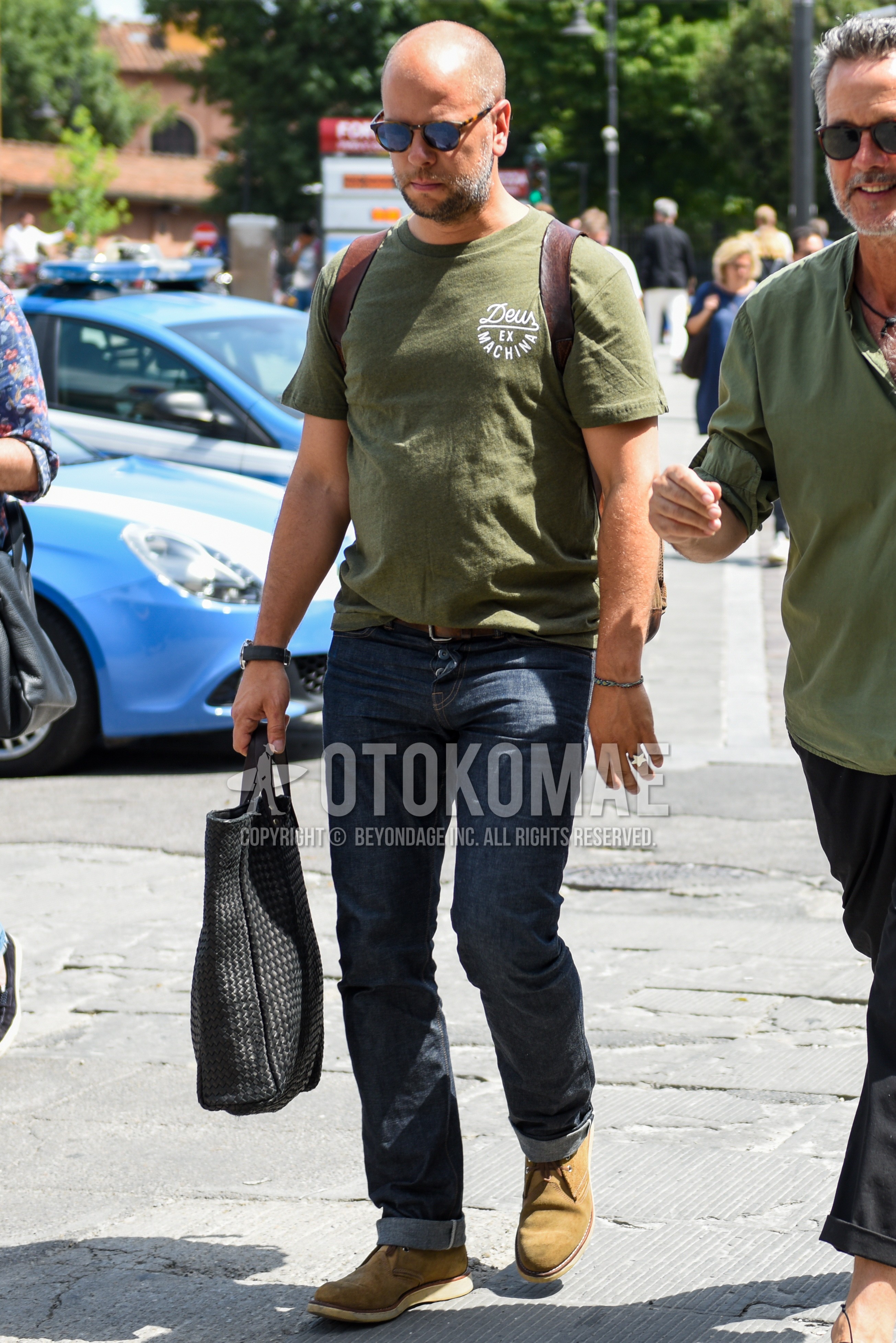 Men's summer outfit with black plain sunglasses, olive green plain t-shirt, navy plain denim/jeans, yellow beige chukka boots, black plain tote bag.