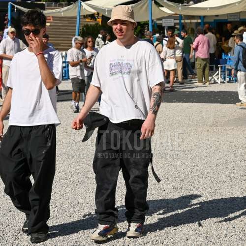 ベージュ無地のバケットハット、黒レタードのベースボールキャップ、白グラフィックのTシャツ、ブラウン無地のメッシュベルト、黒ボトムスのチノパン、マルチカラーローカットのスニーカーを合わせた夏春のメンズコーデ・着こなし。