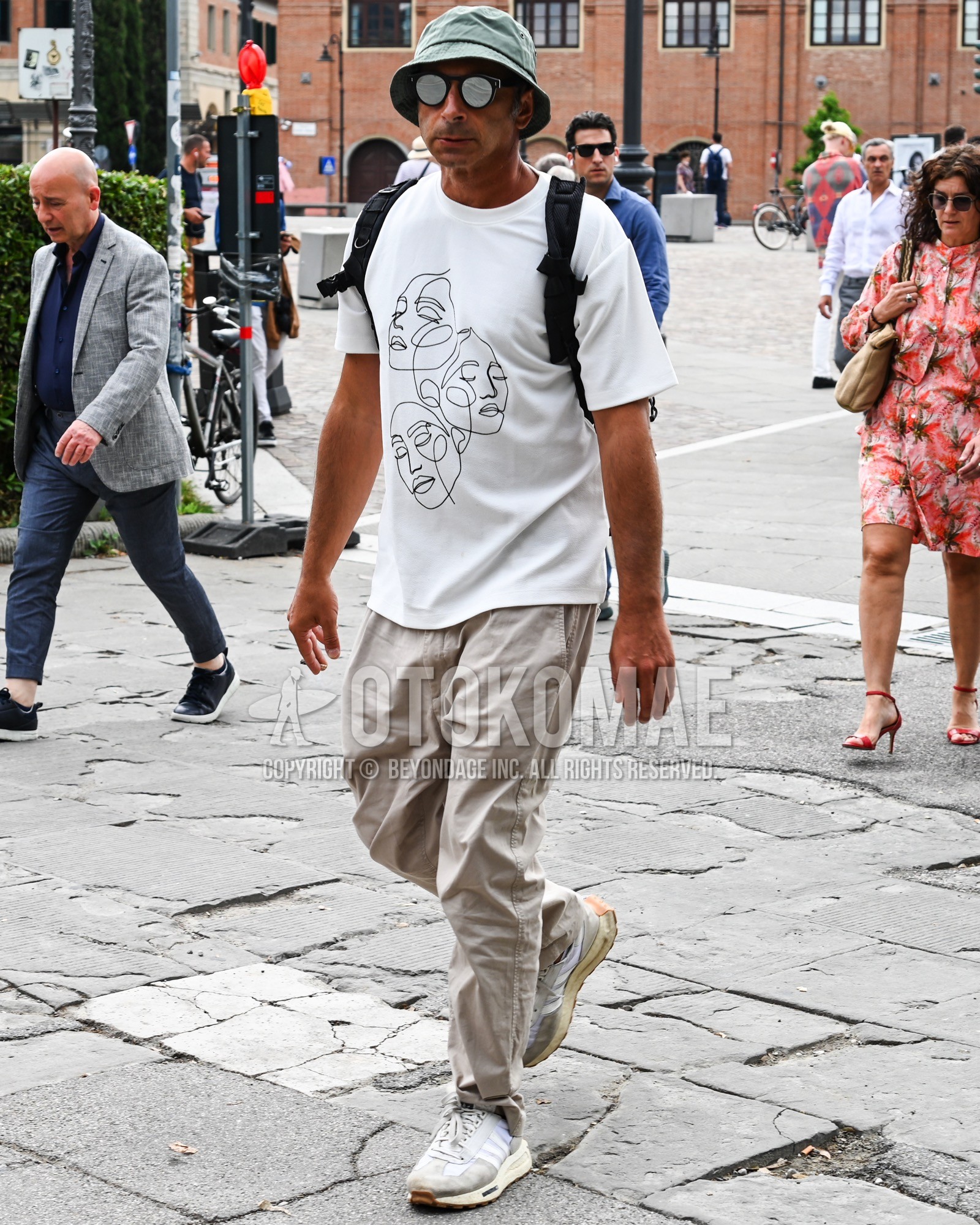 Men's spring summer outfit with green plain bucket hat, silver plain sunglasses, white tops/innerwear t-shirt, beige plain chinos, white low-cut sneakers, black plain backpack.