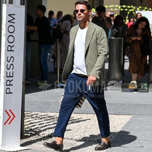 黒・シルバー無地のサングラス、オリーブグリーン無地のテーラードジャケット、白無地のTシャツ、黒無地のレザーベルト、ブルー無地のスラックス、ブラウン・ゴールドコインローファーの革靴、ネイビー・白ストライプのクラッチバッグ/セカンドバッグ/巾着を合わせた秋春のメンズコーデ・着こなし。