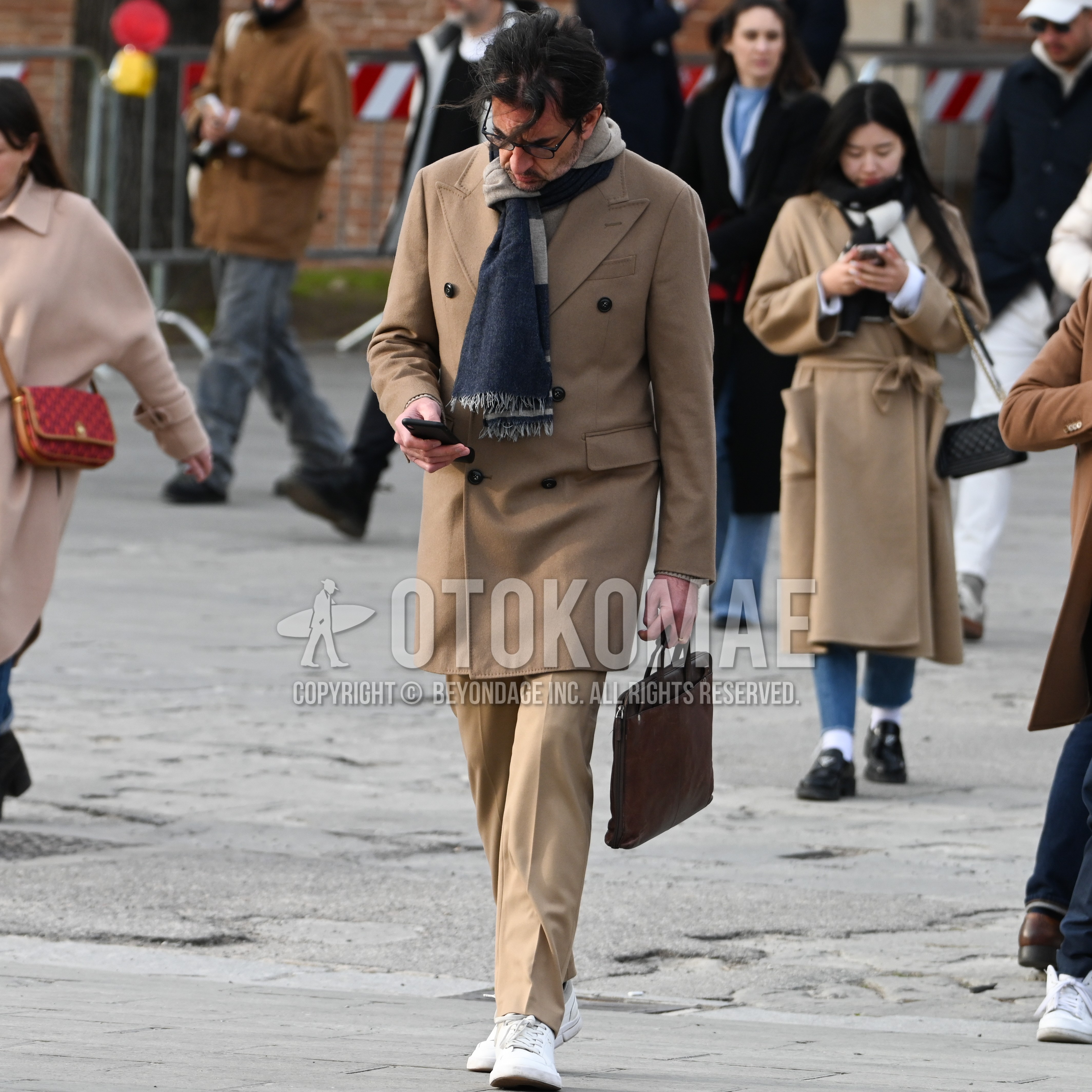 Men's autumn winter outfit with navy beige scarf scarf, beige plain p coat, beige plain slacks, white low-cut sneakers, brown plain briefcase/handbag.
