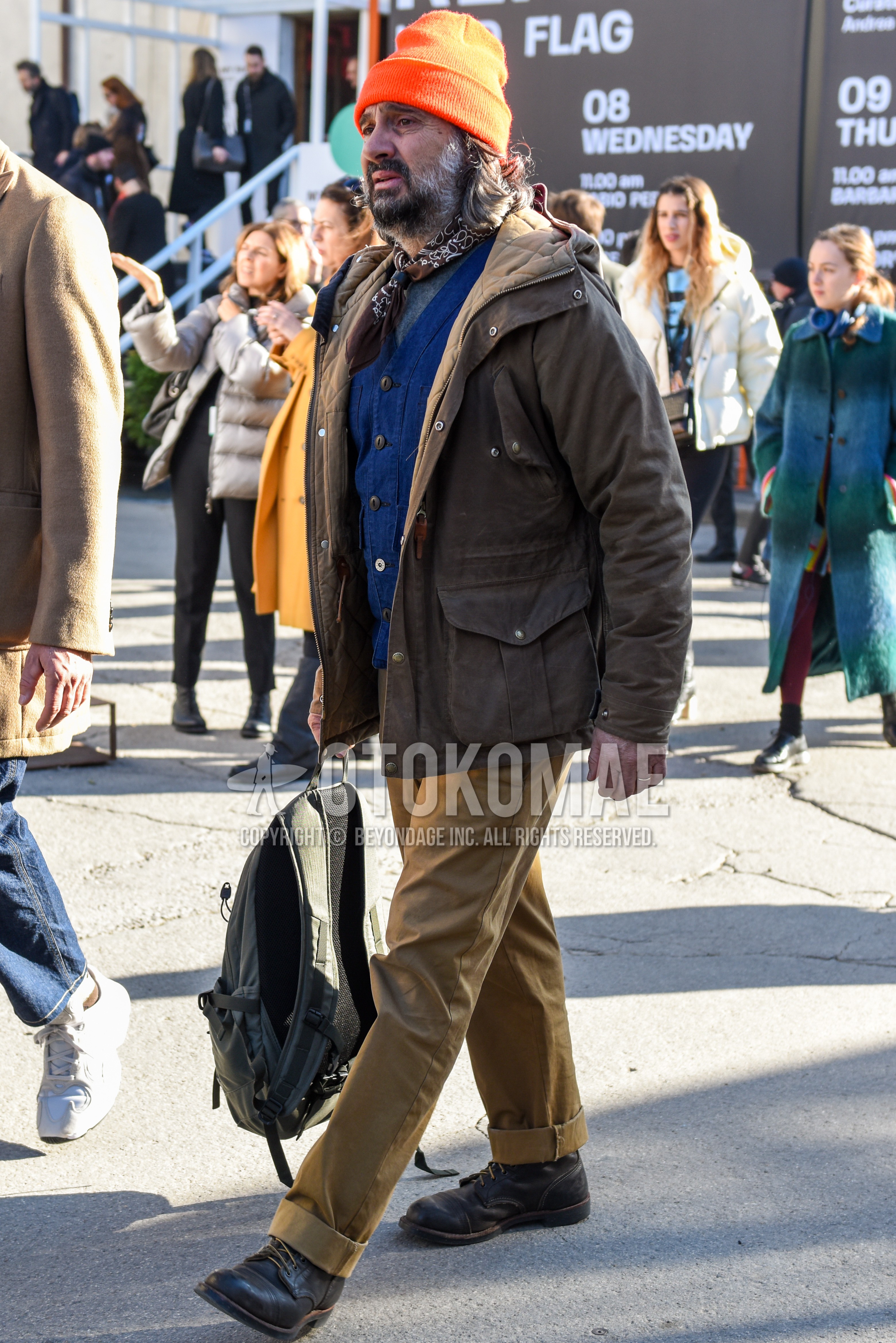 Men's autumn winter outfit with orange plain knit cap, brown paisley bandana/neckerchief, beige plain hooded coat, gray plain t-shirt, blue plain gilet, beige plain chinos, brown  boots, gray plain backpack.