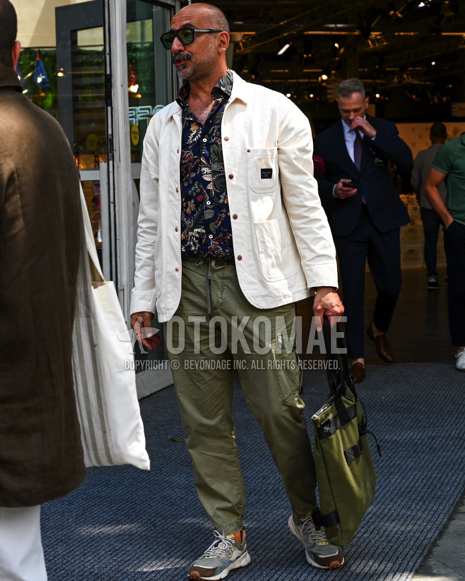 Men's spring summer autumn outfit with black plain sunglasses, white plain coverall, black whole pattern shirt, olive green plain cargo pants, gray low-cut sneakers, olive green plain tote bag.