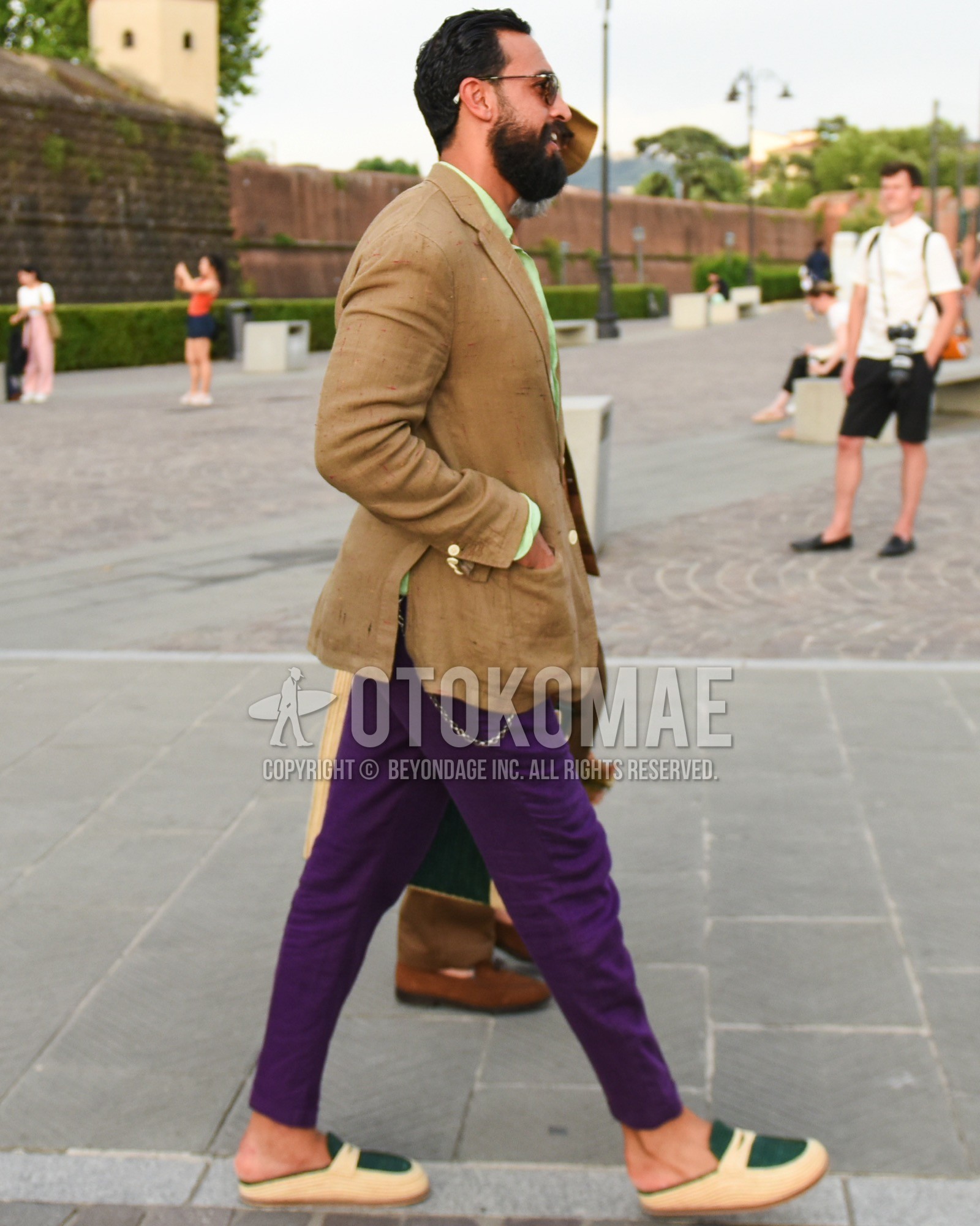 Men's spring summer outfit with brown tortoiseshell sunglasses, beige plain tailored jacket, green plain shirt, purple plain chinos, beige green slip-on sneakers.