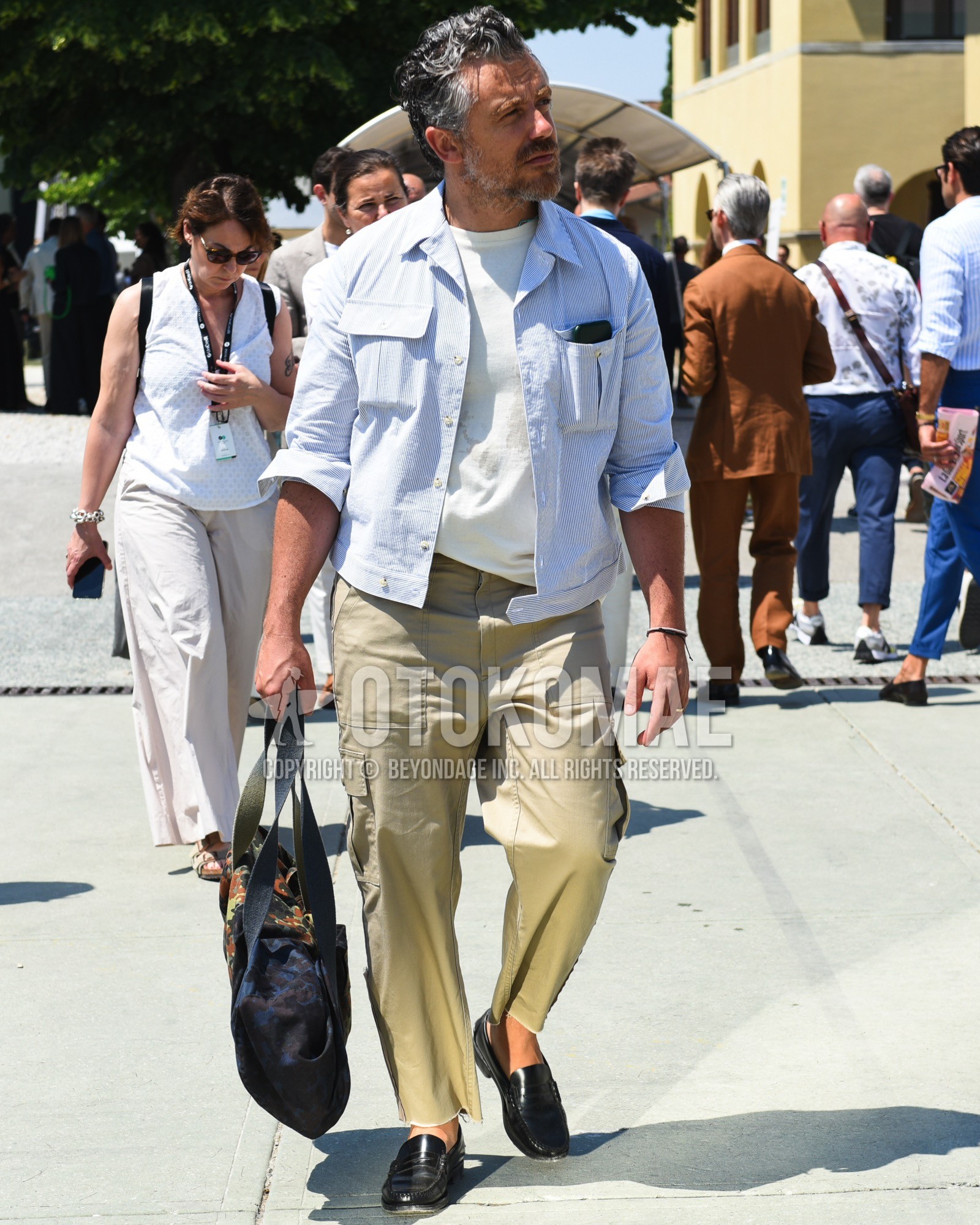 Men's spring summer outfit with blue stripes shirt, white plain t-shirt, beige plain cargo pants, black coin loafers leather shoes, navy bag tote bag.