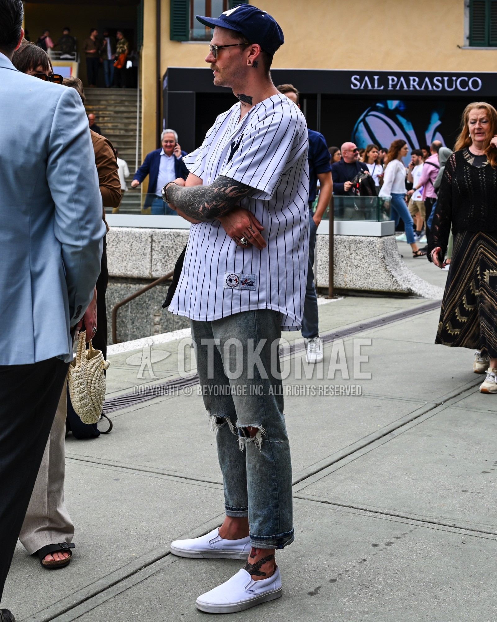 Men's spring summer outfit with navy one point baseball cap, white stripes shirt, light blue plain damaged jeans, white slip-on sneakers.
