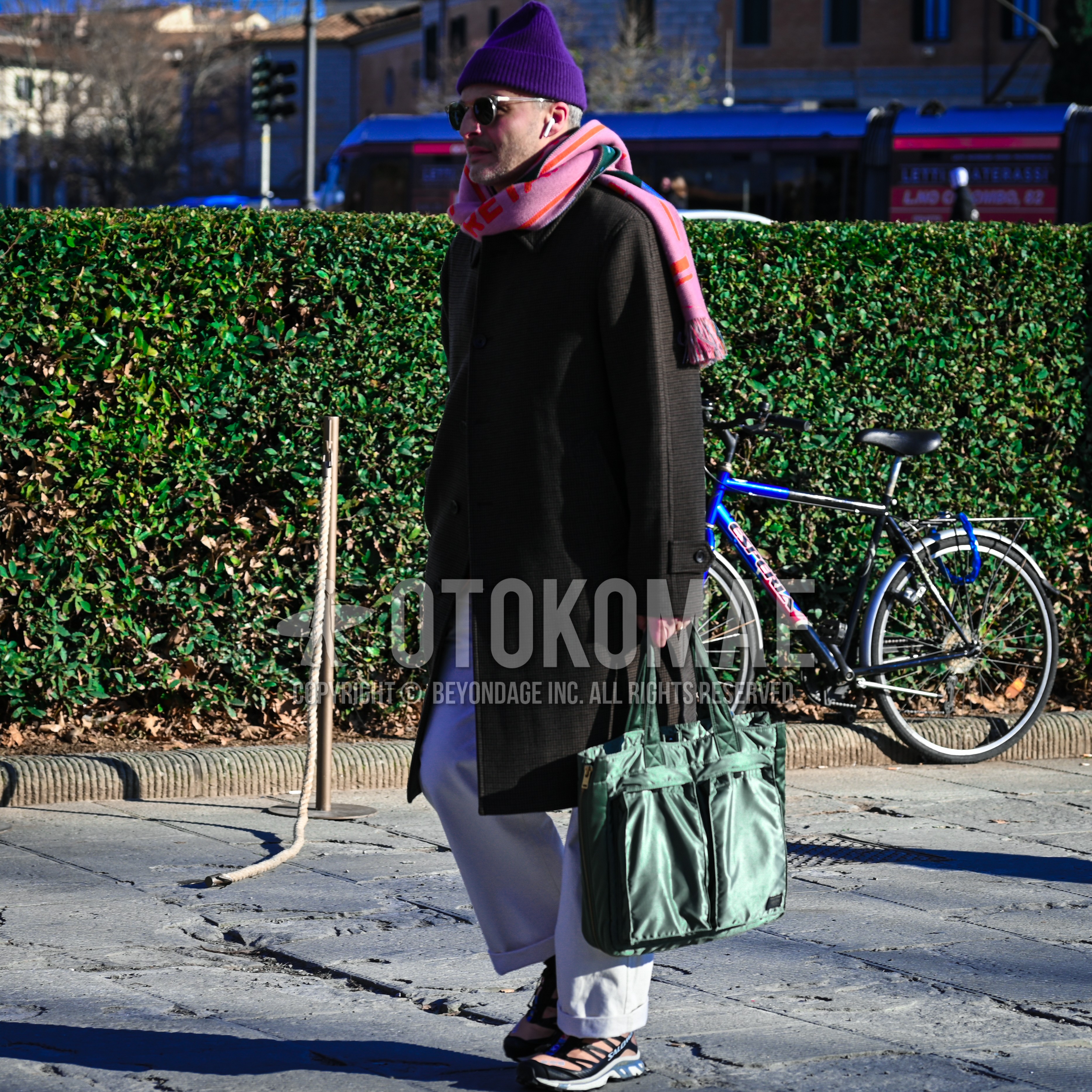 Men's autumn winter outfit with purple plain knit cap, clear plain sunglasses, pink whole pattern scarf, brown check stenkarrer coat, white plain chinos, orange low-cut sneakers, green one point briefcase/handbag.