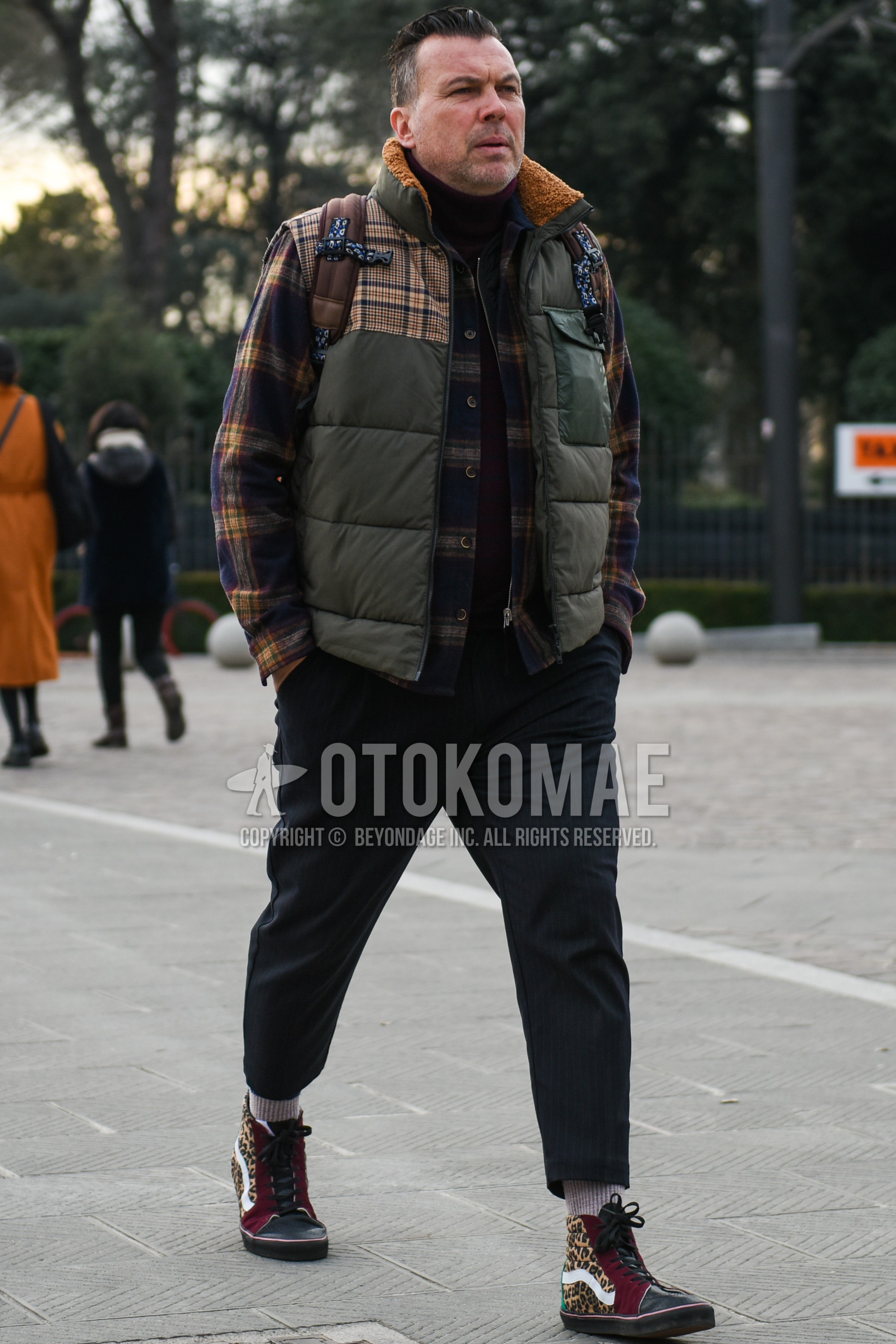 Men's autumn winter outfit with olive green outerwear down jacket, multi-color check shirt, red plain turtleneck knit, olive green tops/innerwear casual vest, navy stripes slacks, navy stripes cropped pants, beige plain socks, multi-color high-cut sneakers.