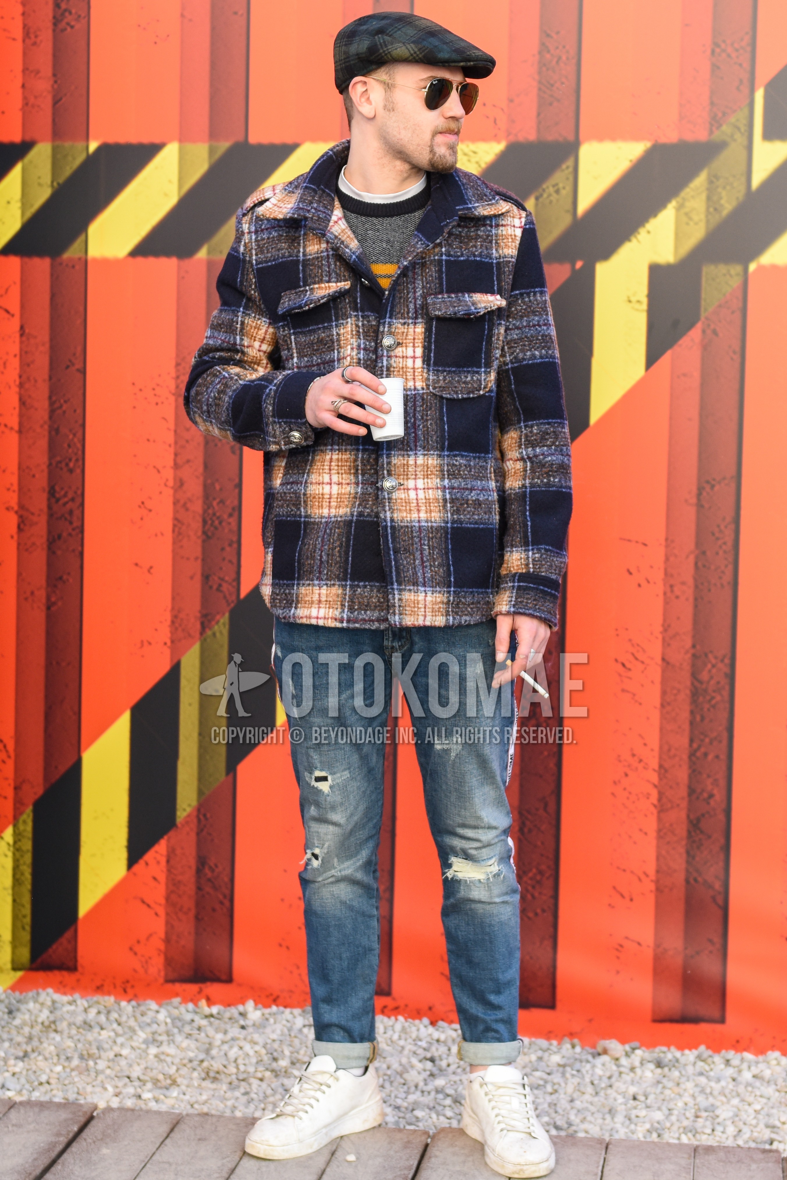 Men's autumn winter outfit with gray check hunting cap, multi-color check shirt jacket, gray graphic sweater, white plain t-shirt, blue plain damaged jeans, white plain socks, white low-cut sneakers.