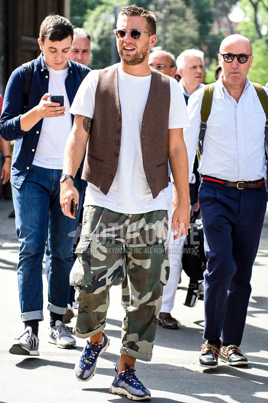 Men's summer outfit with clear plain sunglasses, brown plain gilet, white plain t-shirt, olive green beige brown camouflage cargo pants, clear blue low-cut sneakers.
