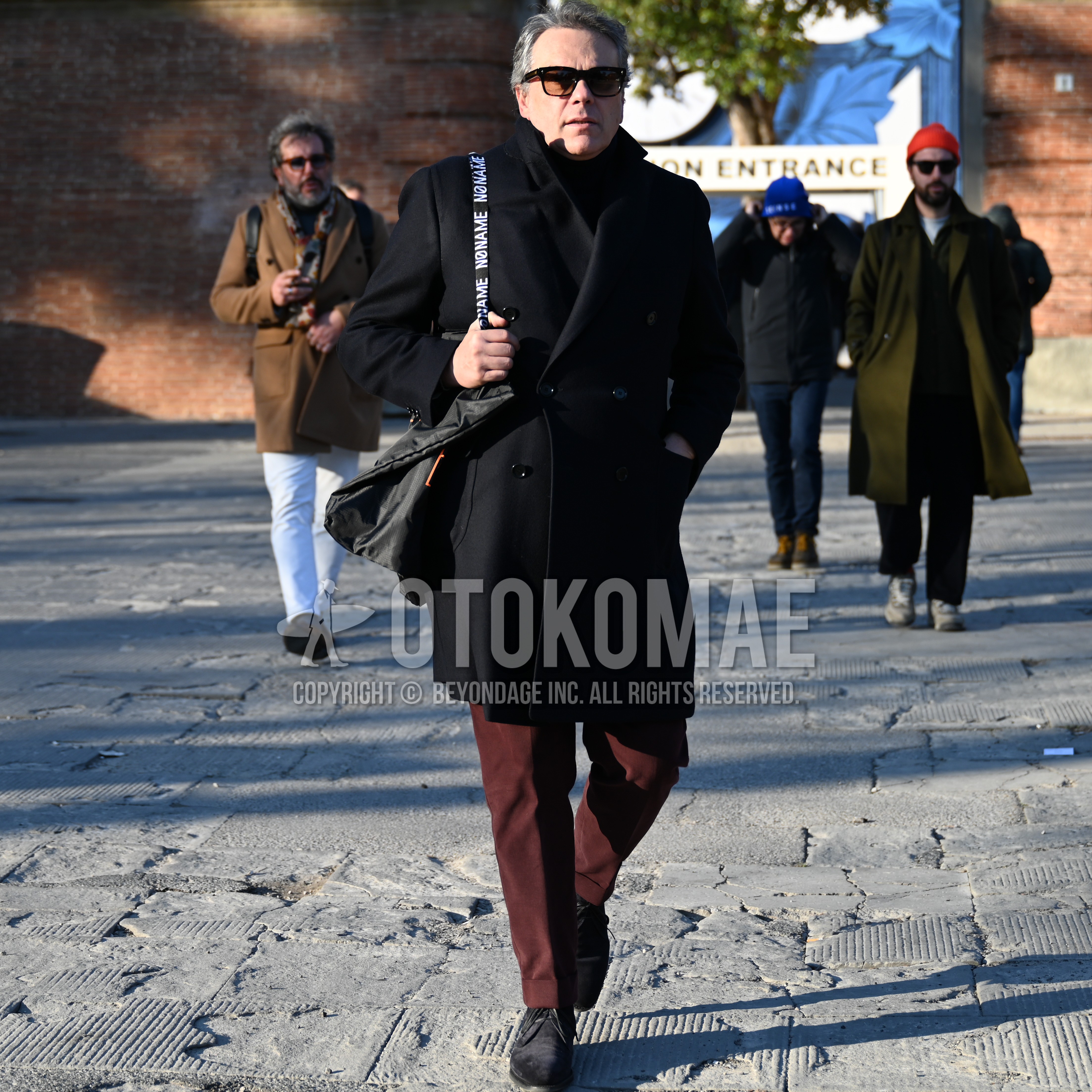 Men's autumn winter outfit with brown tortoiseshell sunglasses, black plain ulster coat, red brown plain slacks, black suede shoes leather shoes, black plain tote bag.