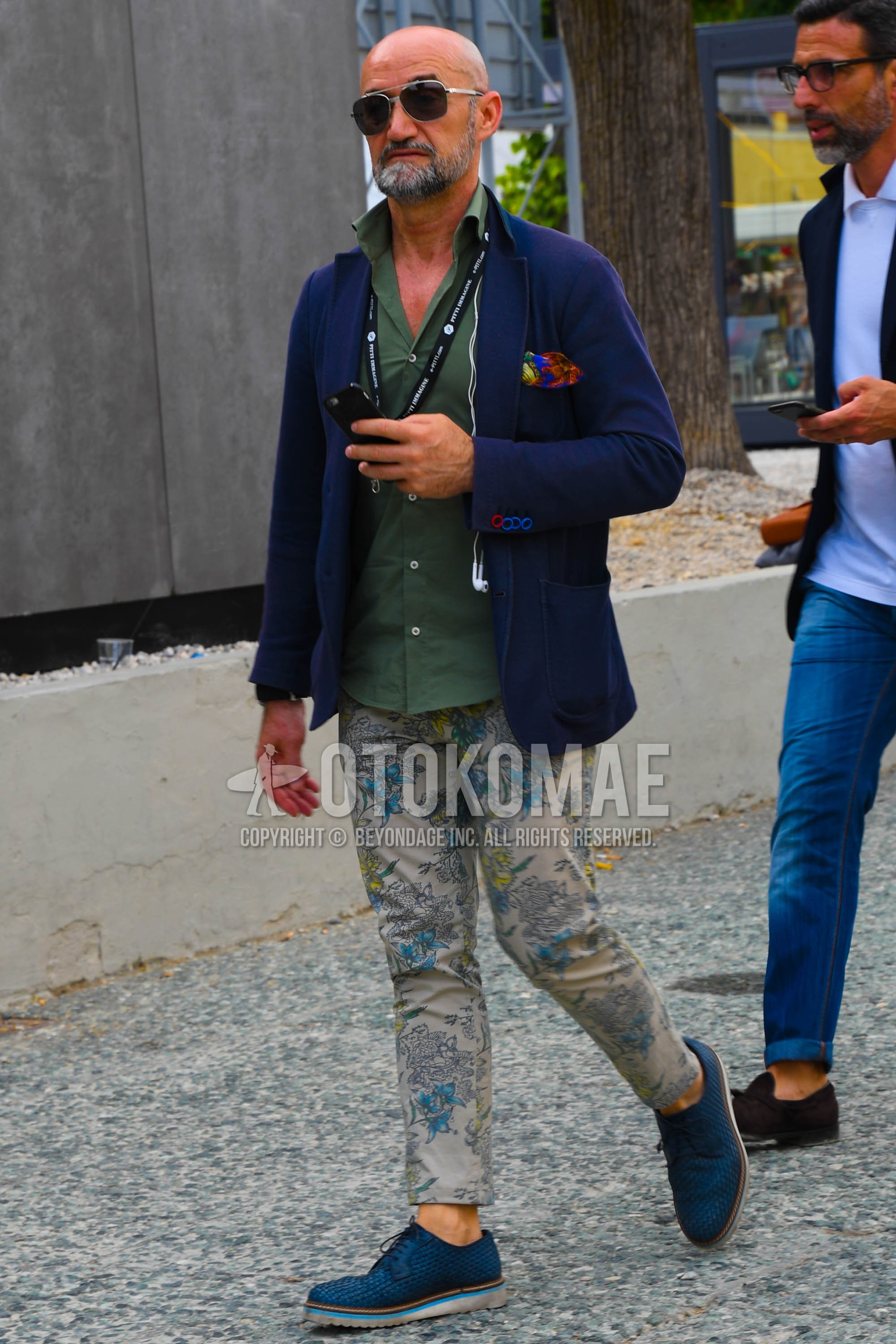 Men's spring summer autumn outfit with silver plain sunglasses, navy plain tailored jacket, green plain shirt, beige bottoms chinos, navy plain toe leather shoes.