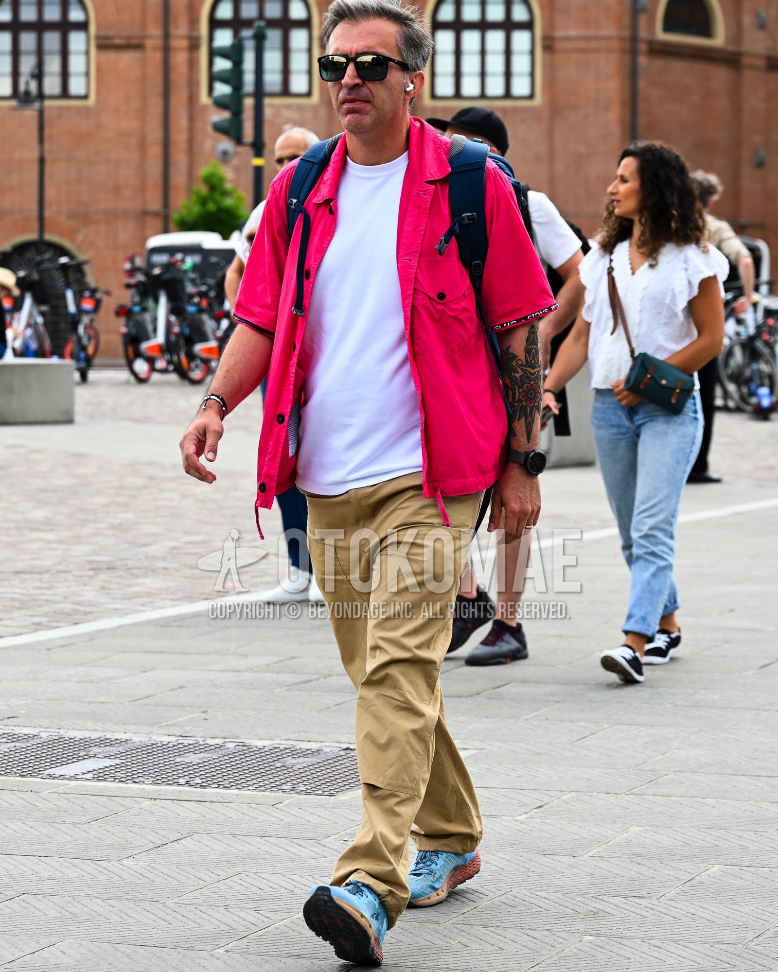 Men's spring summer outfit with black plain sunglasses, pink red plain shirt, white plain t-shirt, beige plain chinos, light blue low-cut sneakers.