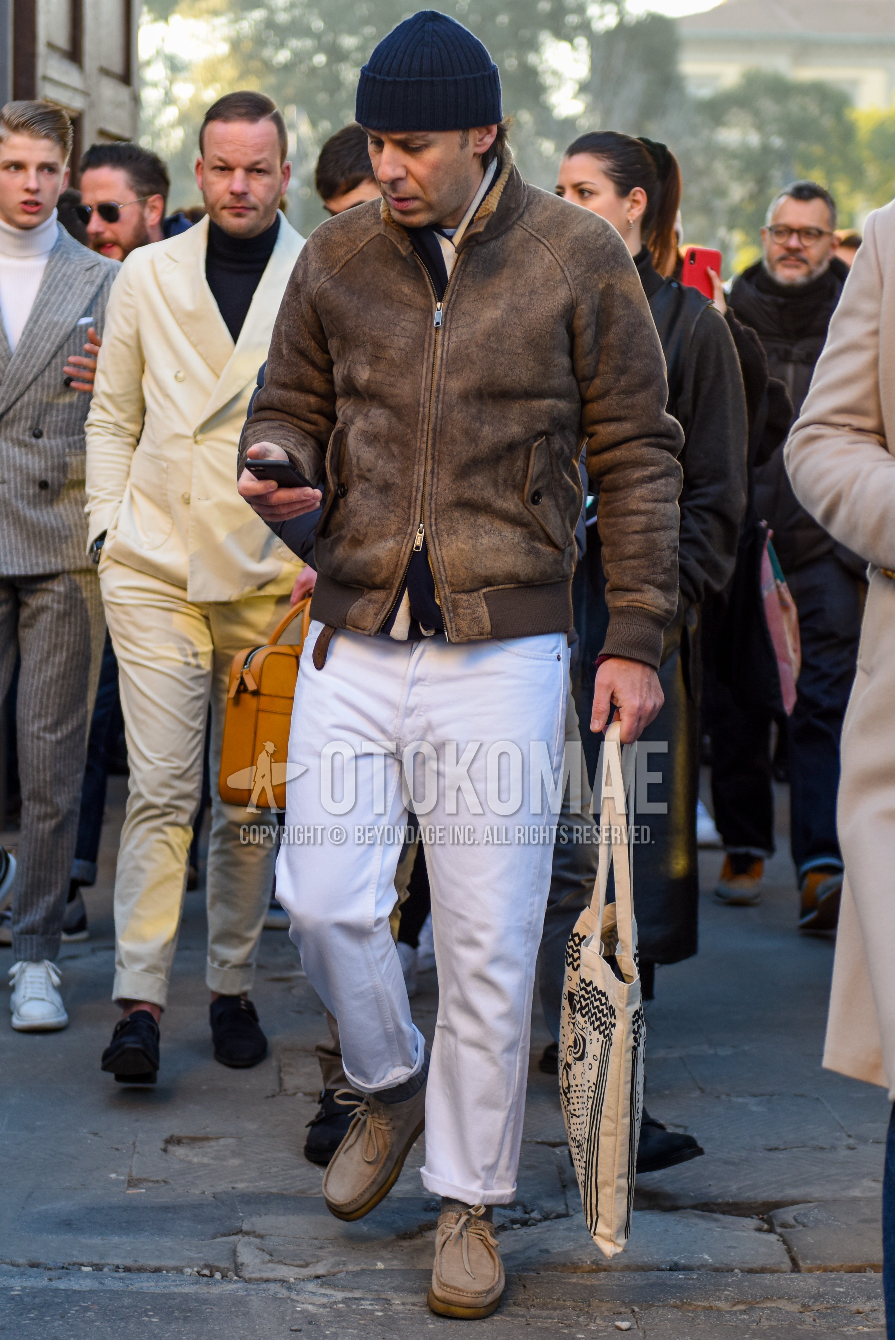 Men's spring autumn outfit with navy plain knit cap, brown plain leather jacket, brown plain military jacket, white plain cotton pants, beige  boots, beige graphic tote bag.