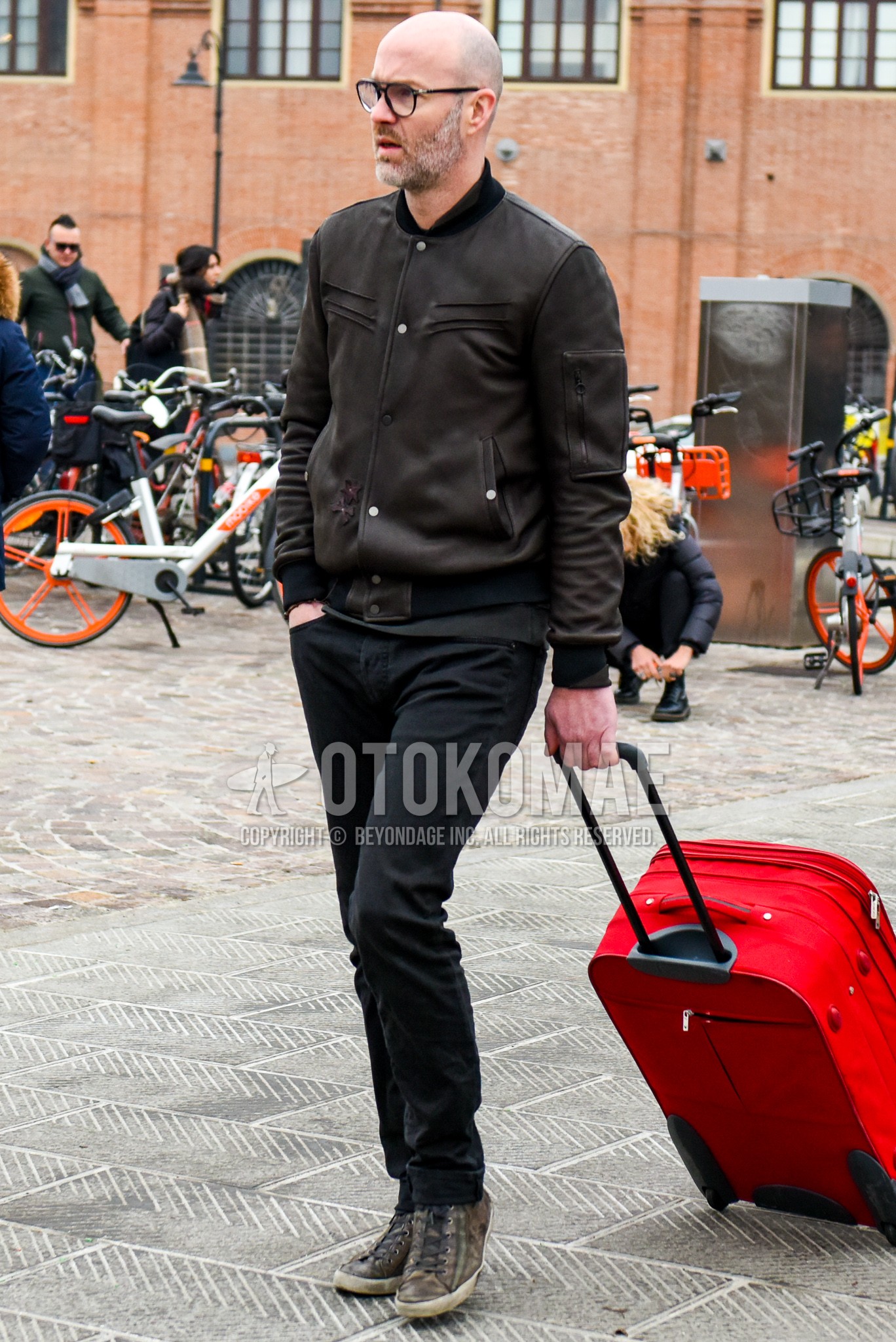 Men's spring autumn winter outfit with black plain glasses, black plain leather jacket, black plain denim/jeans, brown high-cut sneakers, red plain suitcase.
