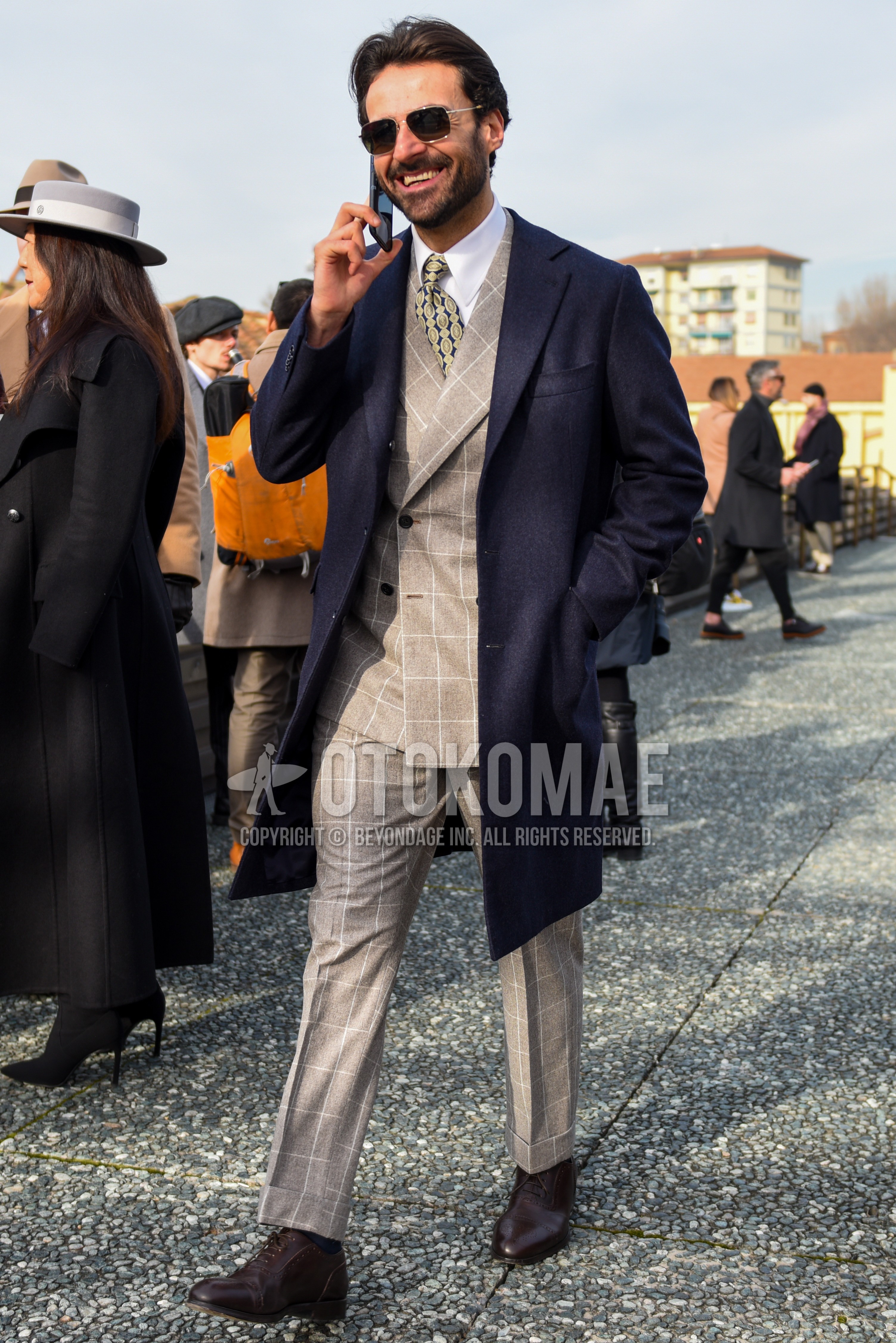 Men's autumn winter outfit with silver plain sunglasses, navy plain chester coat, white plain shirt, brown brogue shoes leather shoes, beige check suit, beige small crest necktie.