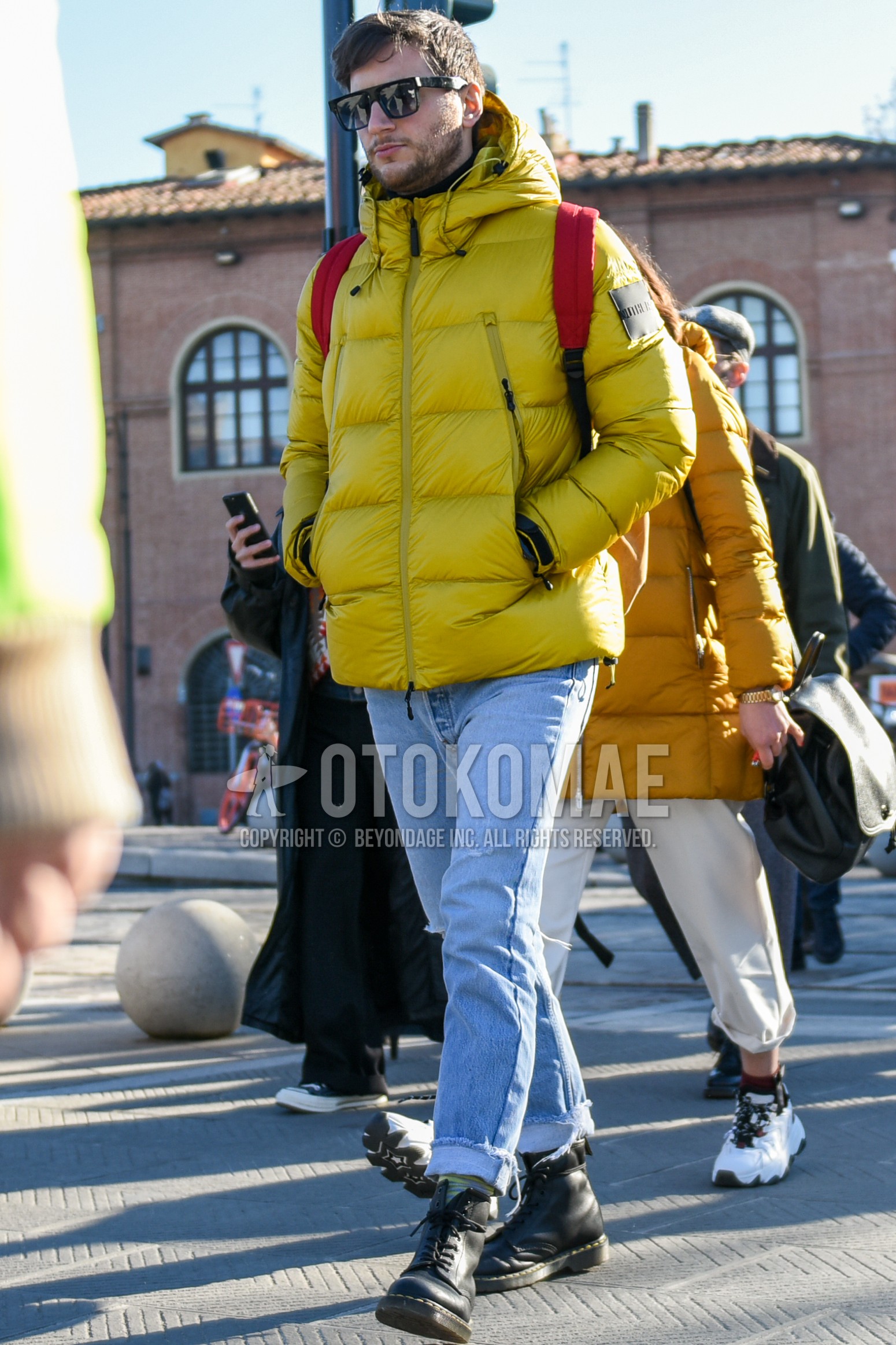 Men's autumn winter outfit with black plain sunglasses, yellow plain down jacket, light blue plain denim/jeans, black  boots, red plain backpack.