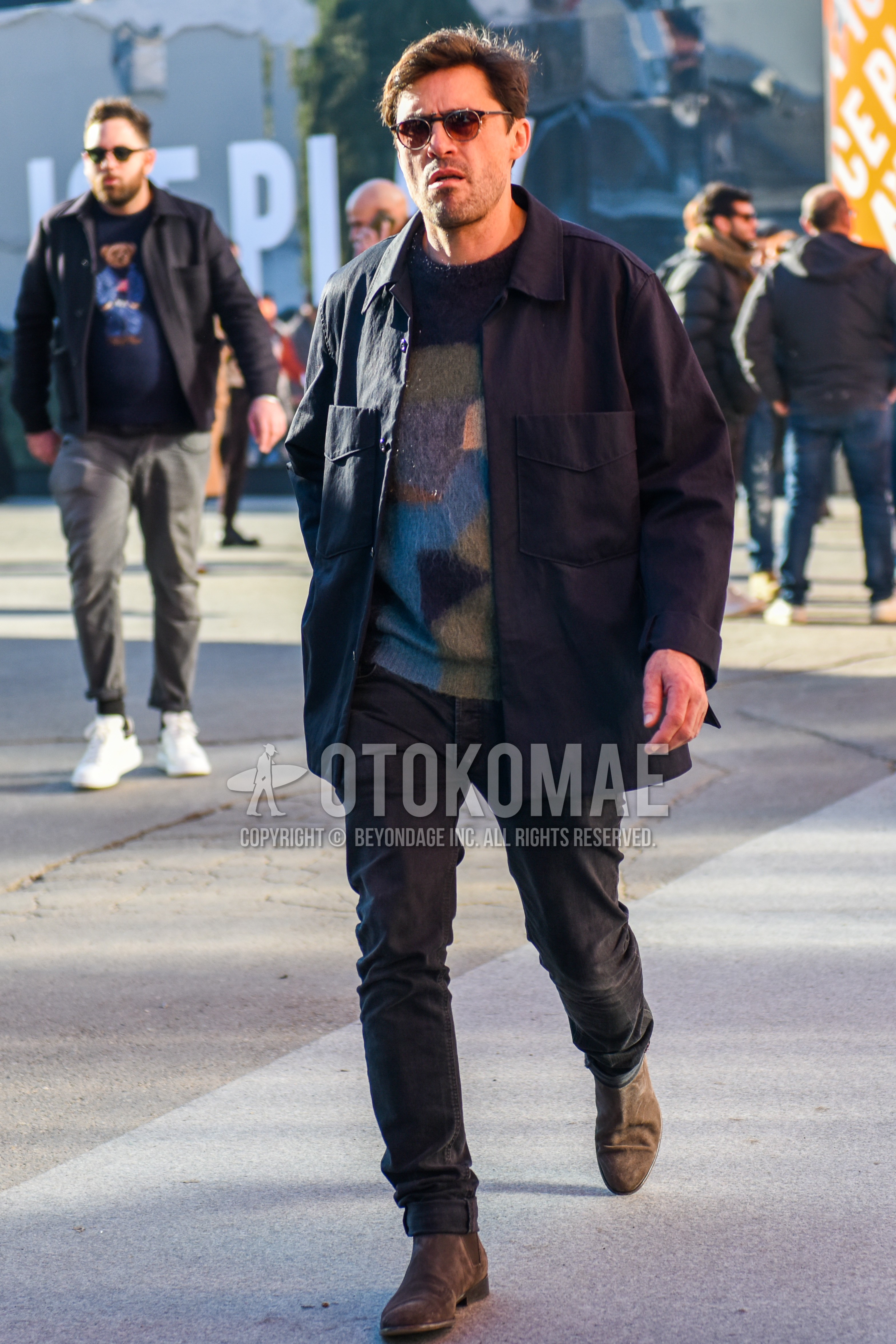 Men's winter outfit with plain sunglasses, dark gray plain shirt jacket, multi-color plain sweater, dark gray plain denim/jeans, brown side-gore boots.