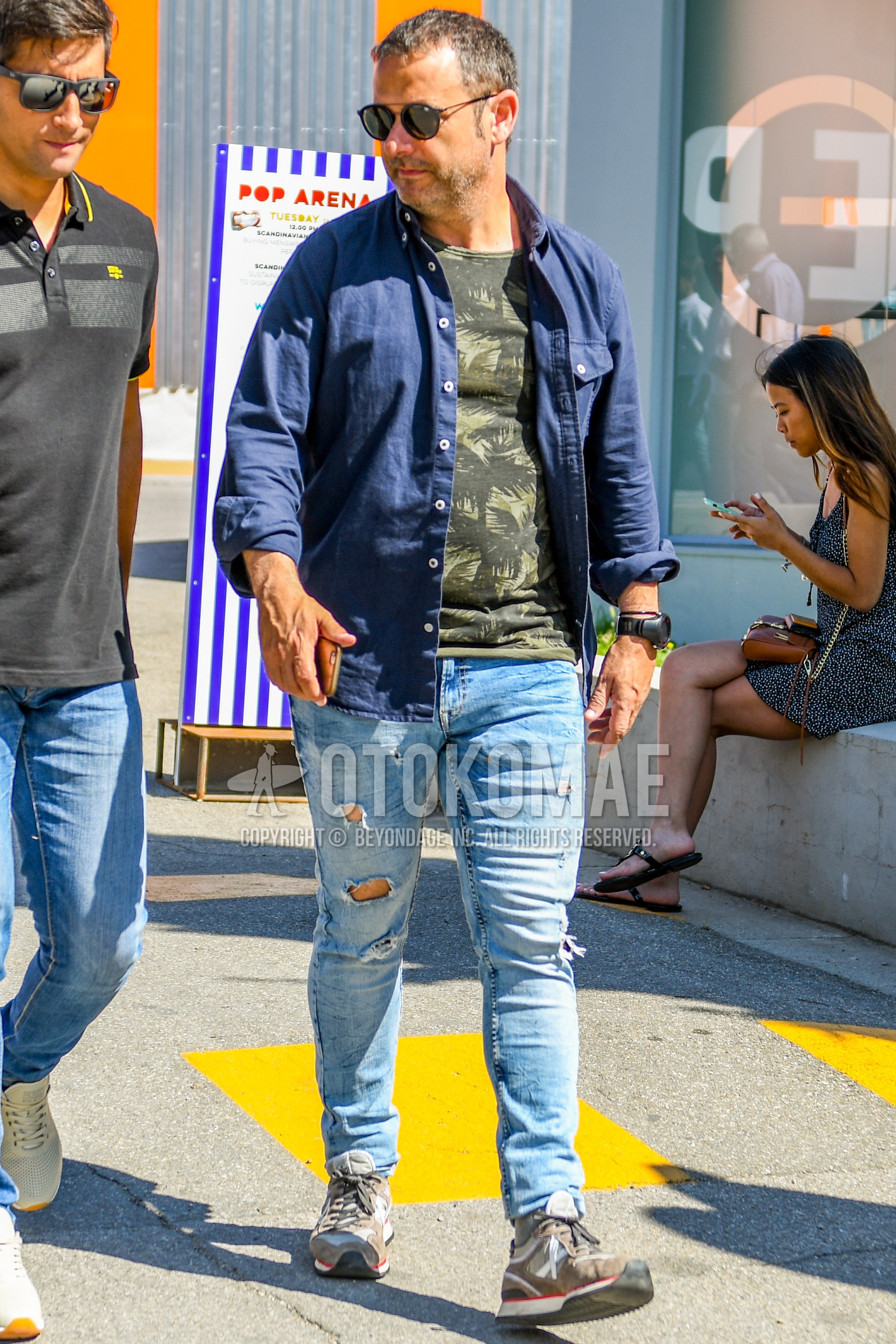 Men's spring summer outfit with plain sunglasses, navy plain shirt, olive green tops/innerwear t-shirt, blue plain denim/jeans, gray low-cut sneakers.