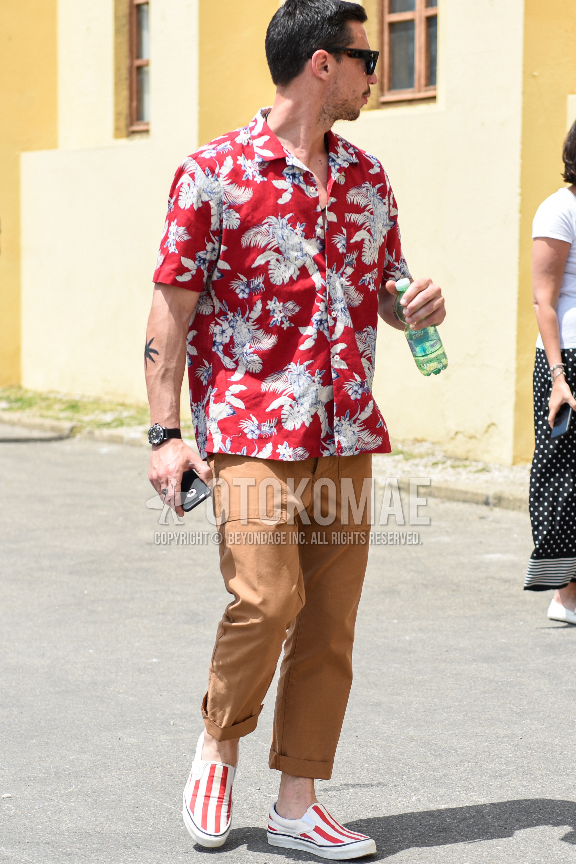 Men's summer outfit with black plain sunglasses, red botanical shirt, brown plain cotton pants, red slip-on sneakers.