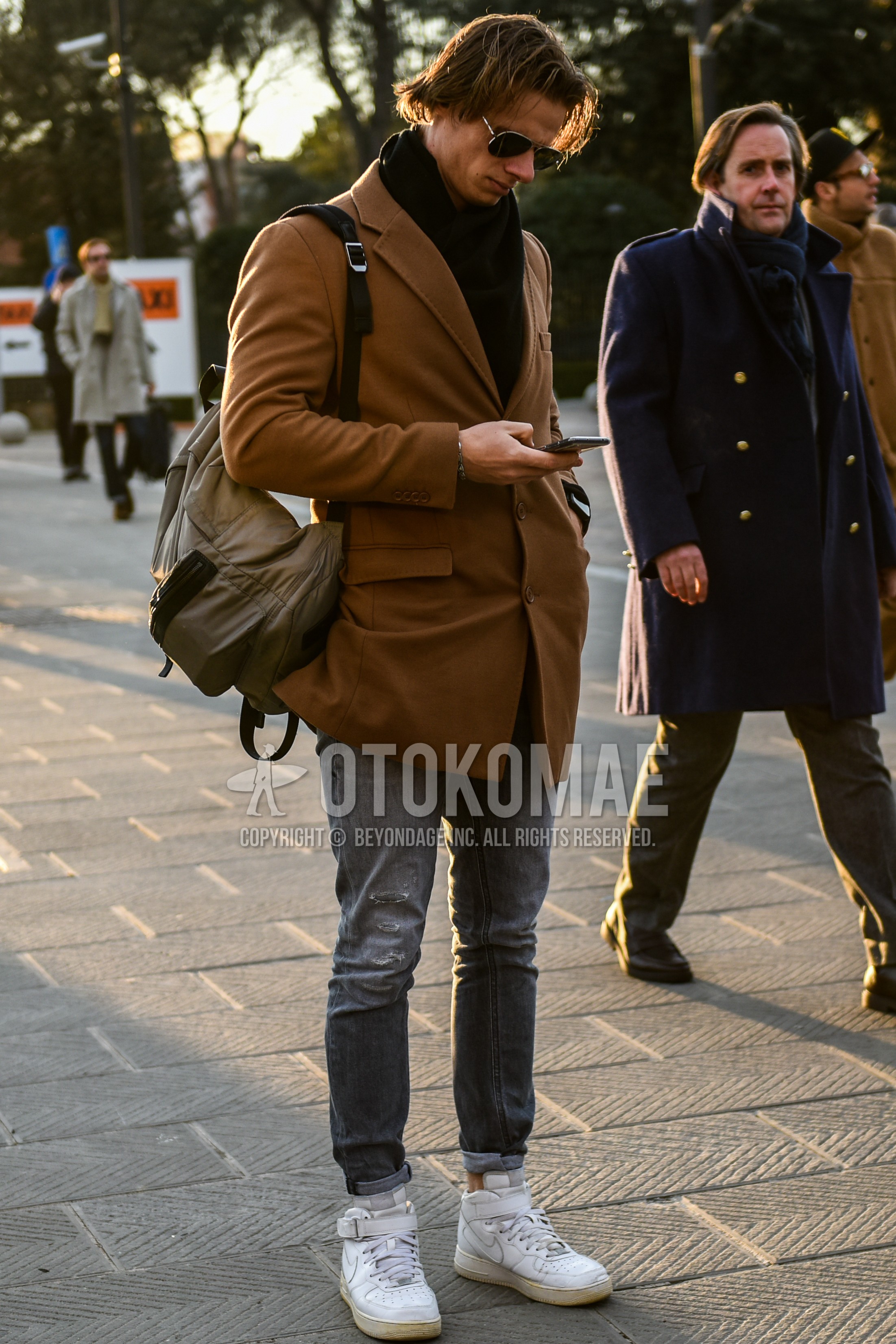 Men's autumn winter outfit with silver plain sunglasses, black plain scarf, beige plain chester coat, gray plain denim/jeans, white high-cut sneakers, beige plain backpack.