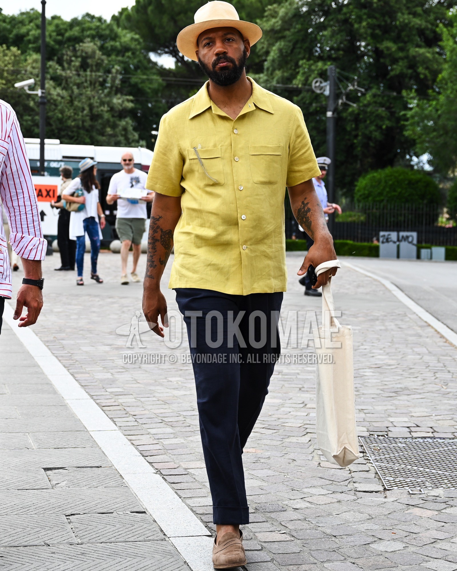 Men's spring summer outfit with beige plain hat, yellow plain shirt, navy plain slacks, brown coin loafers leather shoes, brown suede shoes leather shoes, white plain tote bag.
