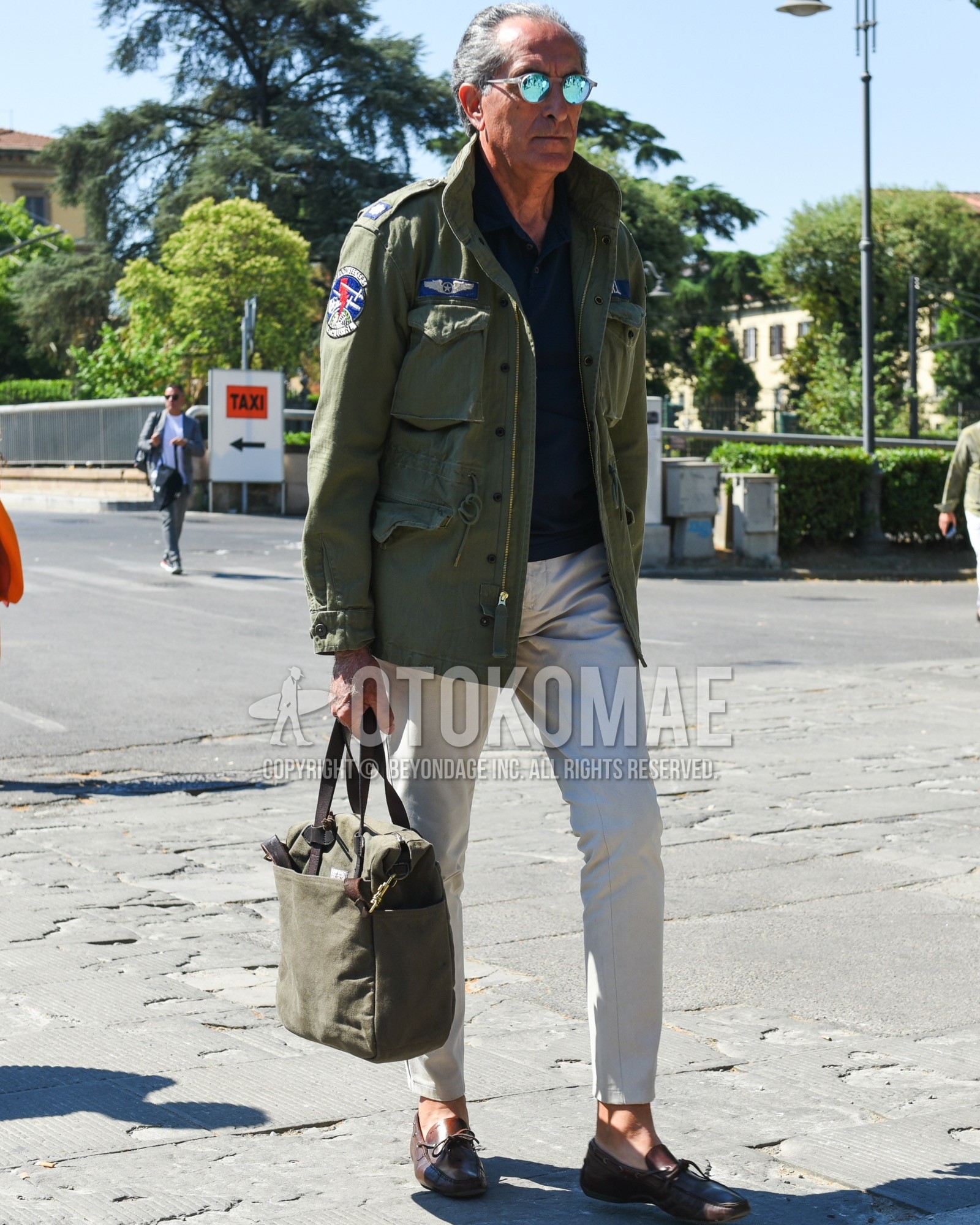 Men's spring summer outfit with silver plain sunglasses, olive green plain field jacket/hunting jacket, navy plain polo shirt, white plain cotton pants, brown moccasins/deck shoes leather shoes, olive green plain tote bag.