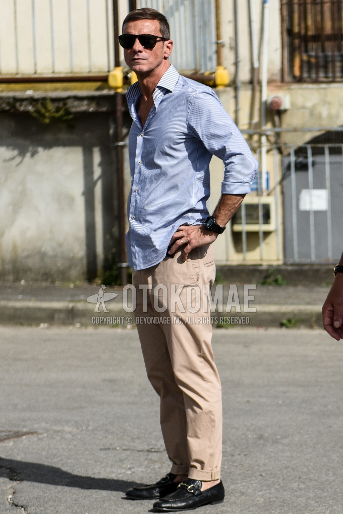 Men's summer outfit with plain sunglasses, light blue stripes shirt, beige plain chinos, black bit loafers leather shoes.