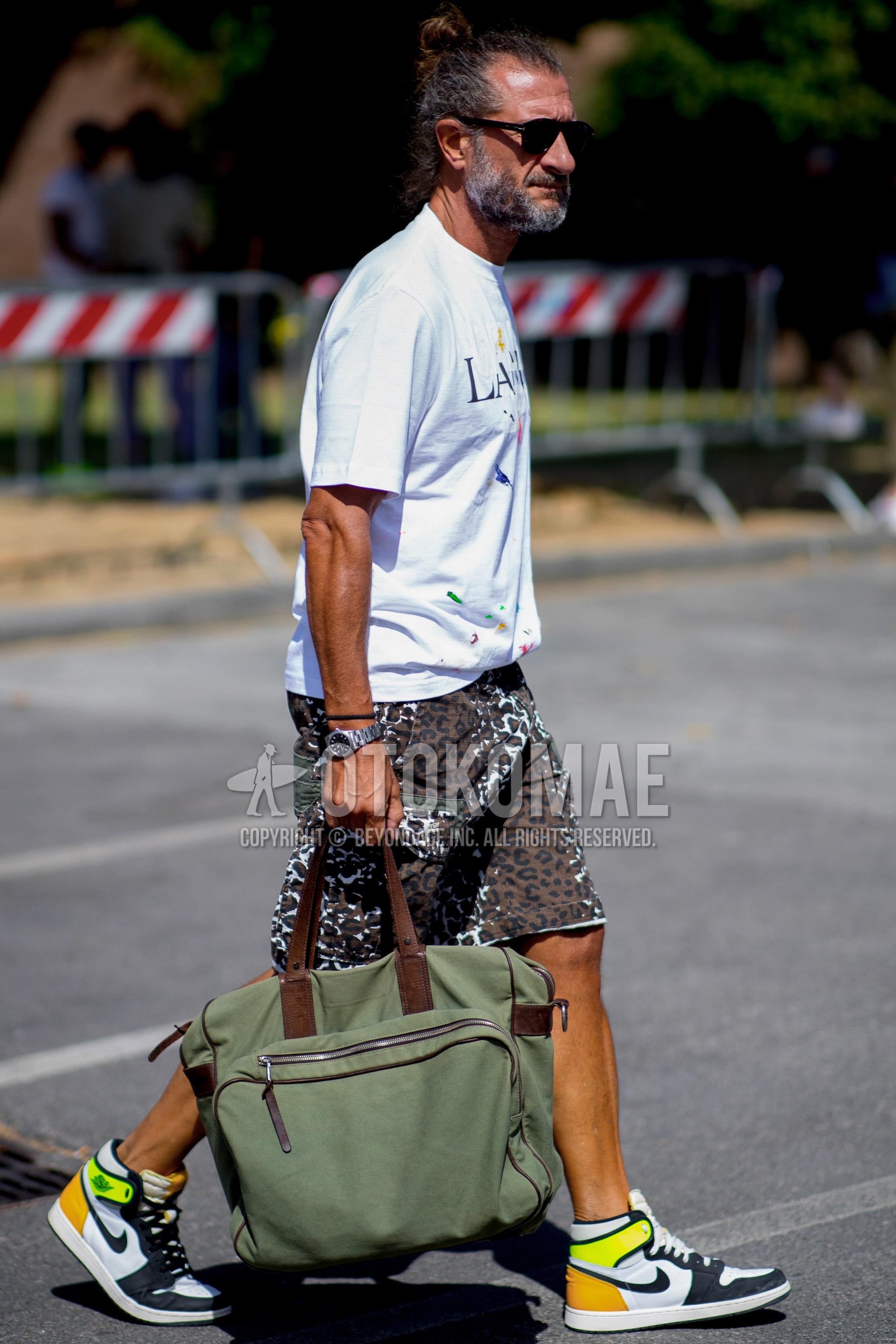 Men's spring summer outfit with black plain sunglasses, white graphic t-shirt, brown leopard short pants, white sneakers, olive green plain tote bag.