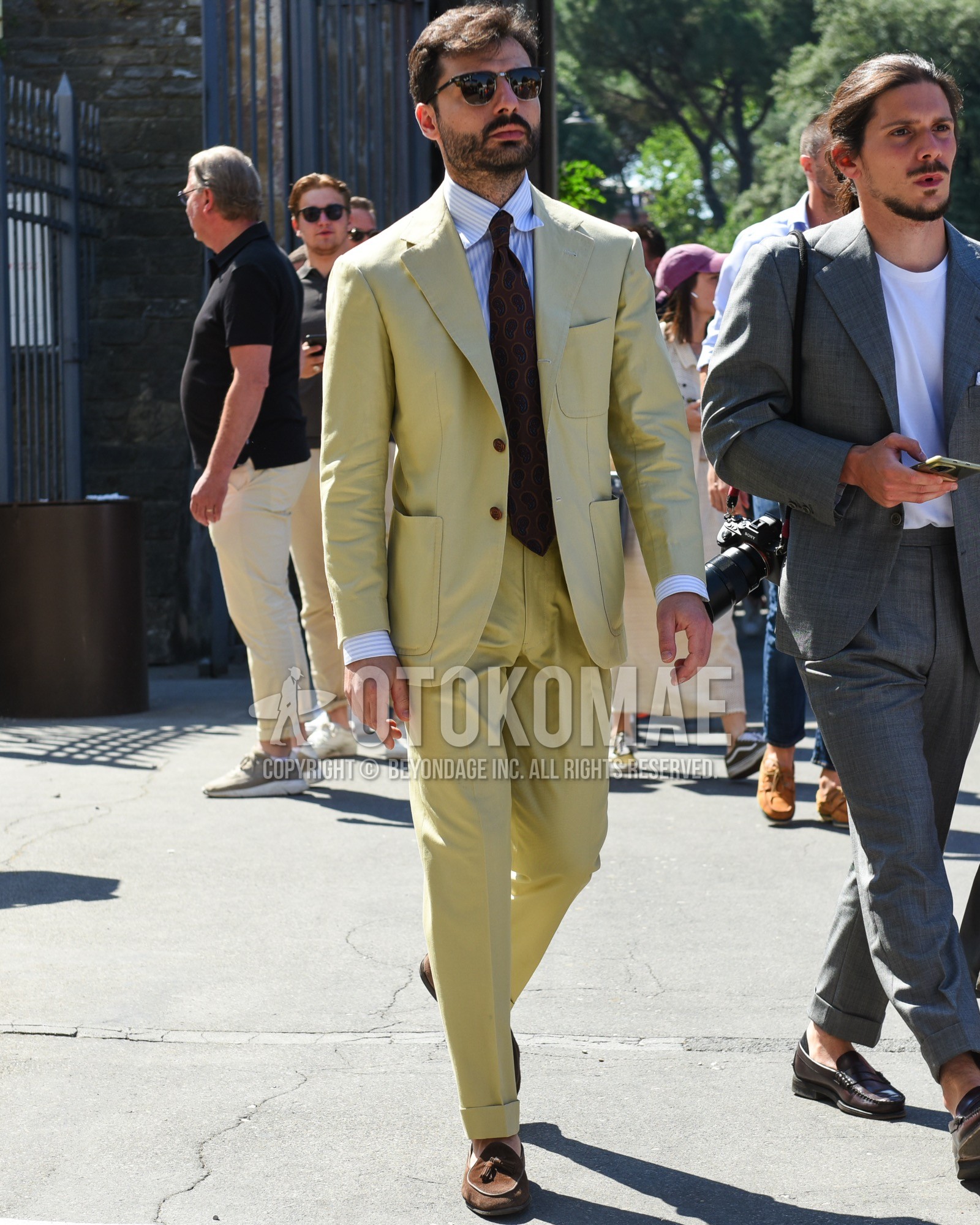 Men's spring summer outfit with black plain sunglasses, white beige stripes shirt, brown tassel loafers leather shoes, brown suede shoes leather shoes, yellow plain suit, brown small crest necktie.