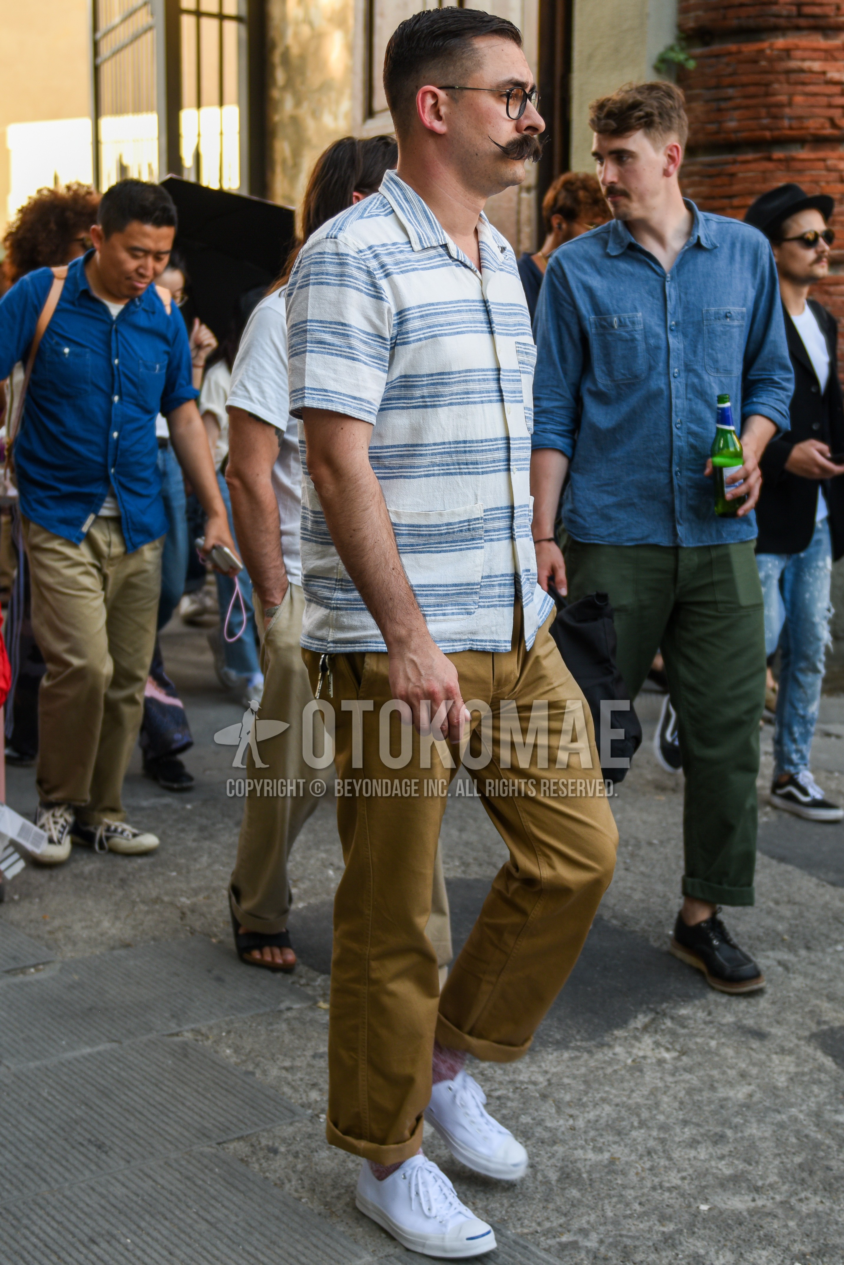 Men's summer outfit with black plain glasses, white light blue horizontal stripes shirt, beige plain chinos, white low-cut sneakers.