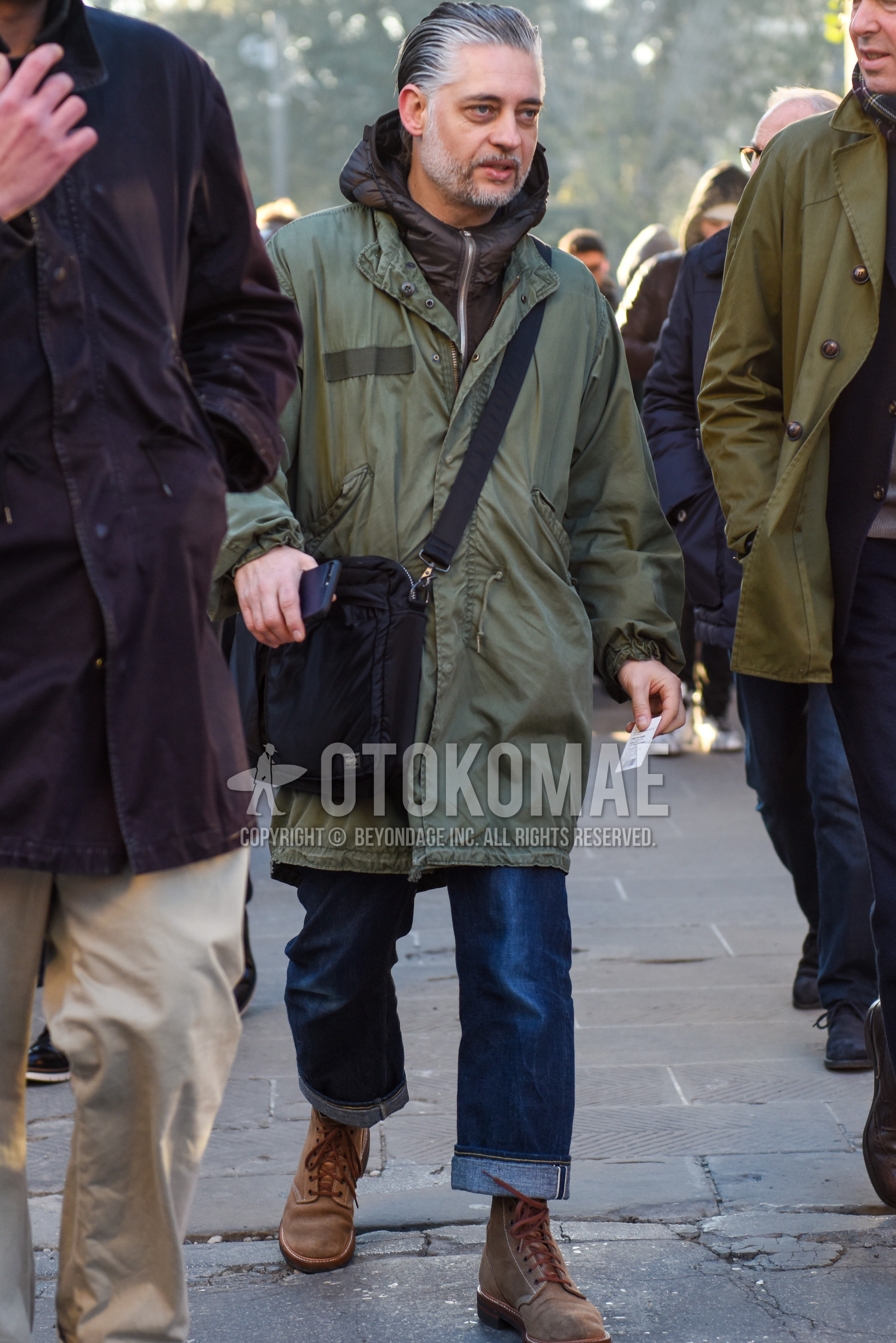 Men's autumn winter outfit with olive green plain mod coat, brown plain hoodie, blue plain denim/jeans, beige work boots, black plain shoulder bag.
