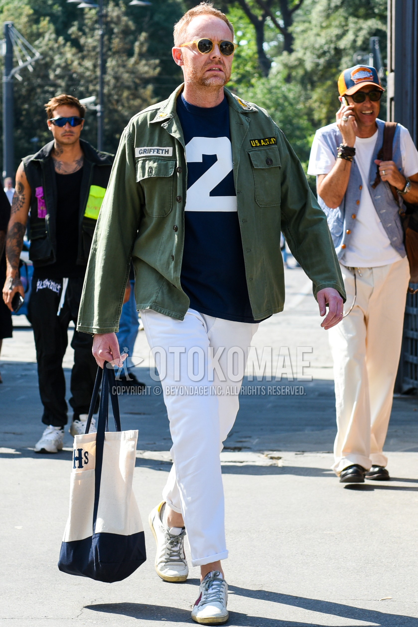 Men's spring autumn outfit with beige plain sunglasses, olive green plain shirt jacket, navy deca logo t-shirt, white plain chinos, white low-cut sneakers, beige one point tote bag.