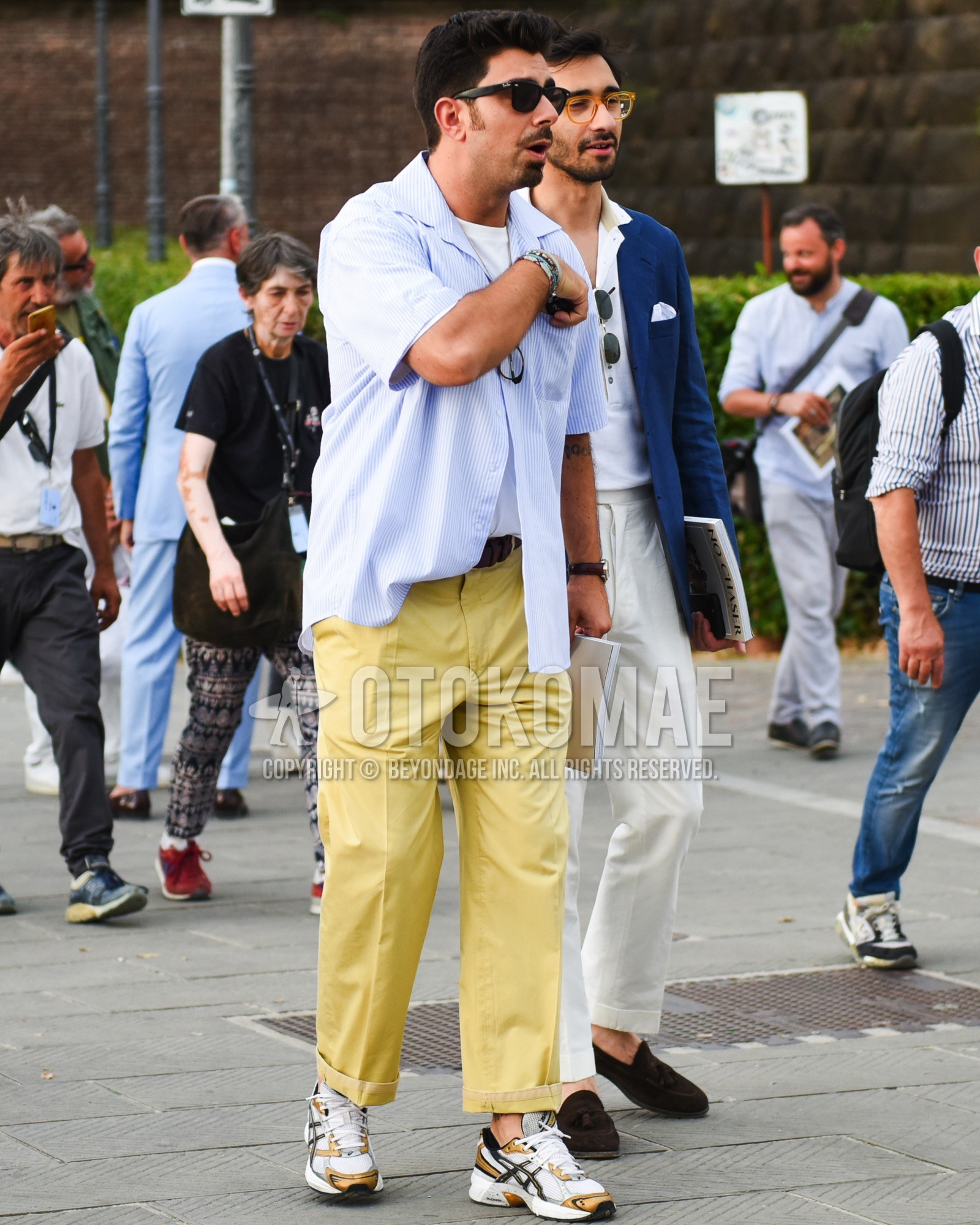 Men's spring summer outfit with black plain sunglasses, blue white stripes shirt, white plain t-shirt, brown plain leather belt, yellow plain slacks, gray low-cut sneakers.