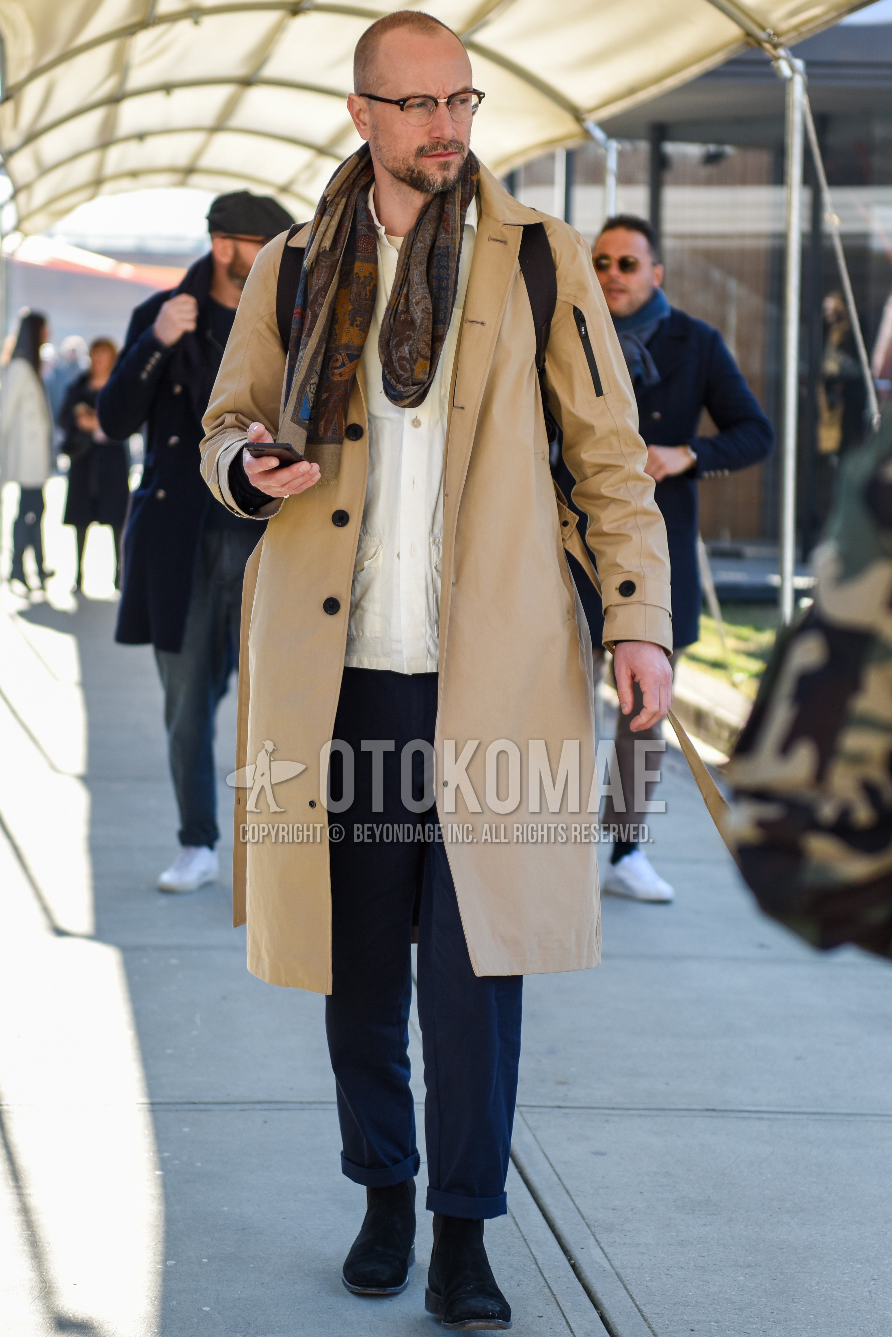 Men's autumn winter outfit with brown tortoiseshell glasses, brown scarf scarf, beige plain stenkarrer coat, white plain shirt, gray plain slacks, black side-gore boots.