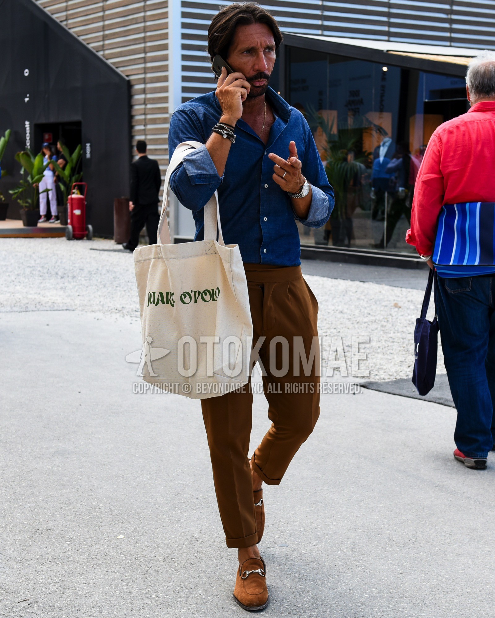Men's spring summer outfit with blue plain denim shirt/chambray shirt, brown plain slacks, brown bit loafers leather shoes, white deca logo tote bag.