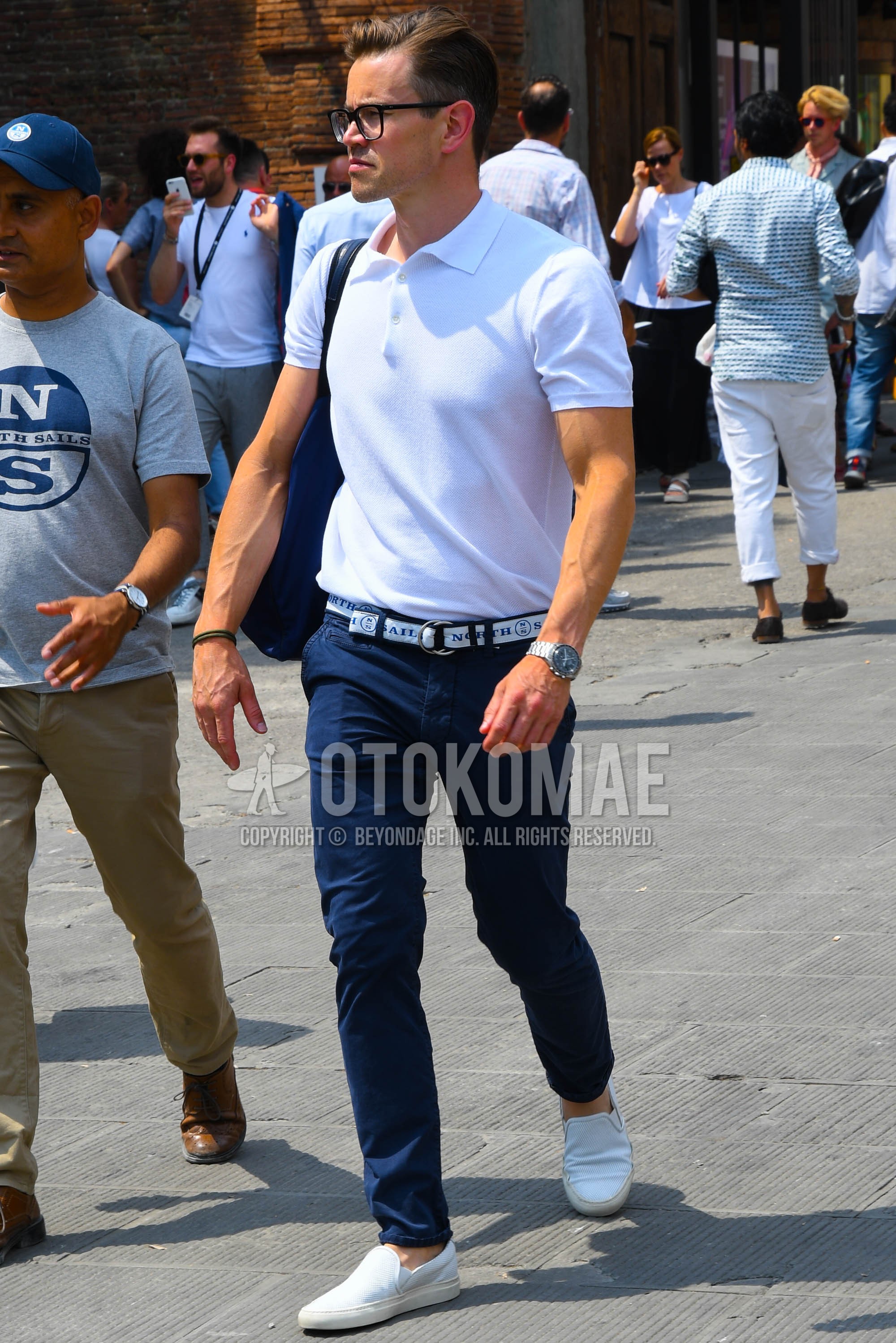 Men's summer outfit with plain glasses, white plain polo shirt, white graphic braided belt, navy plain chinos, white slip-on sneakers.