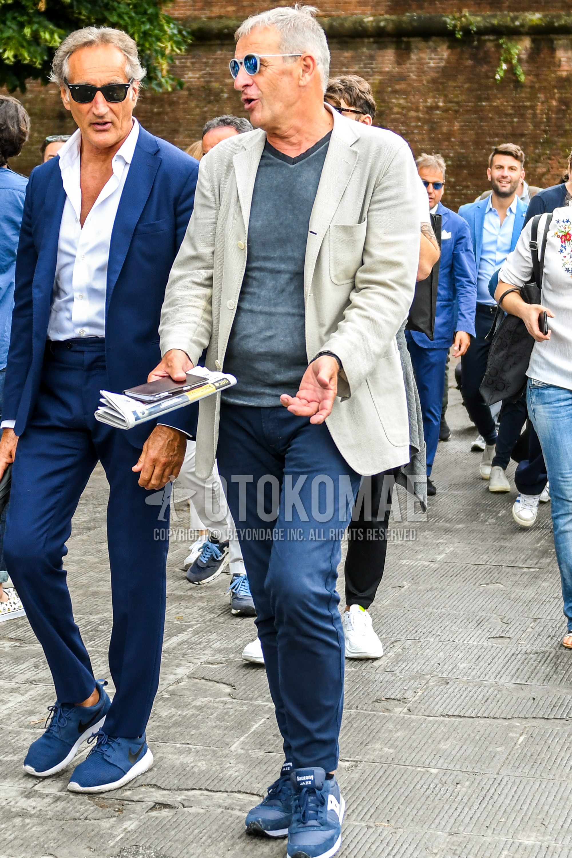 Men's spring summer autumn outfit with gray plain sunglasses, beige plain tailored jacket, gray plain t-shirt, navy plain cotton pants, blue low-cut sneakers.