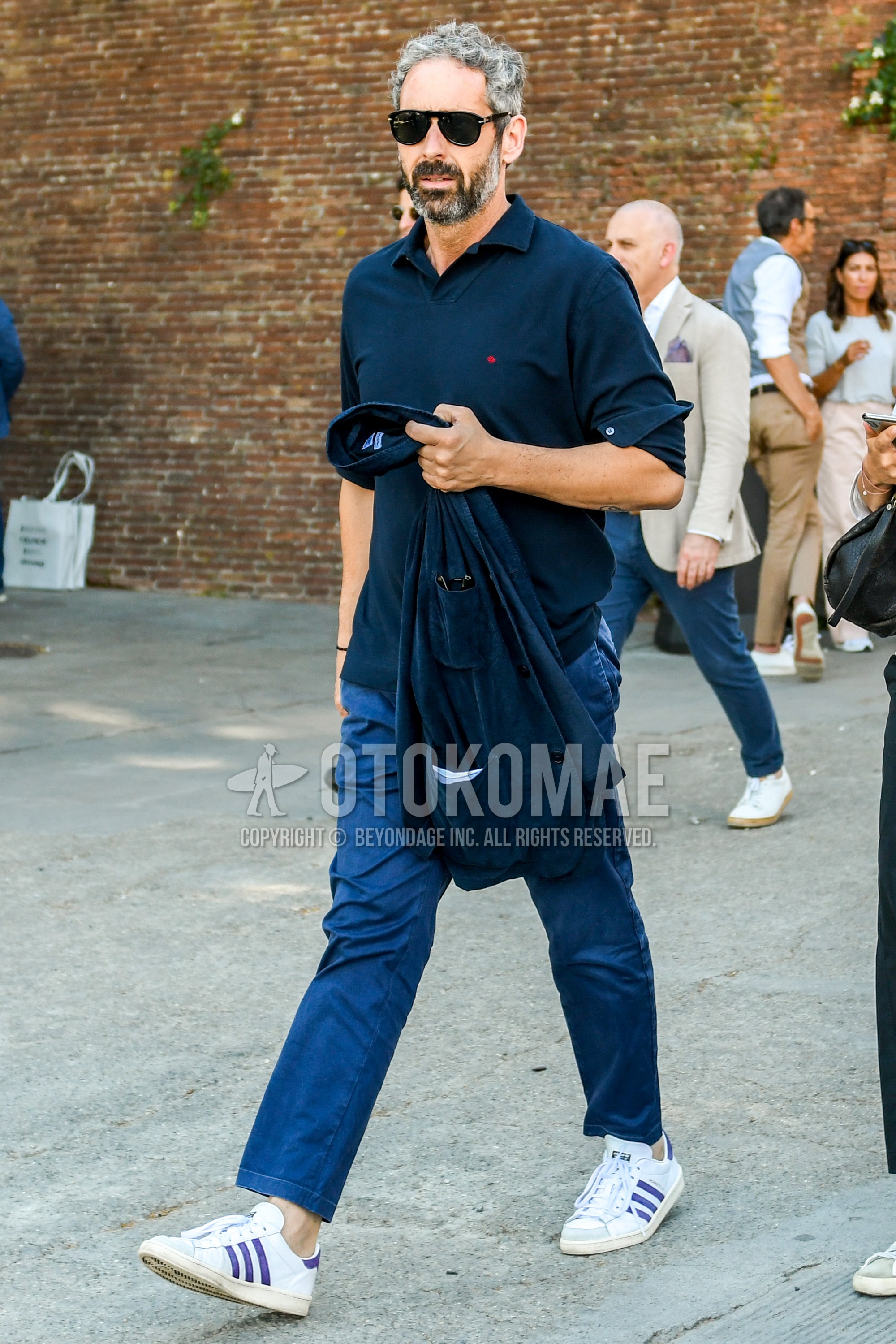 Men's spring summer outfit with black plain sunglasses, navy one point polo shirt, blue plain chinos, blue plain ankle pants, white low-cut sneakers.